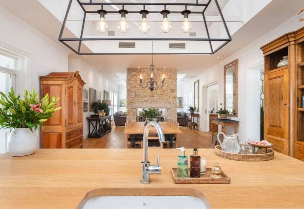 Open kitchen with a wooden countertop, potted flowers, and personal care bottles, overlooking a dining and living area featuring a stone fireplace, wooden furniture, and large windows.