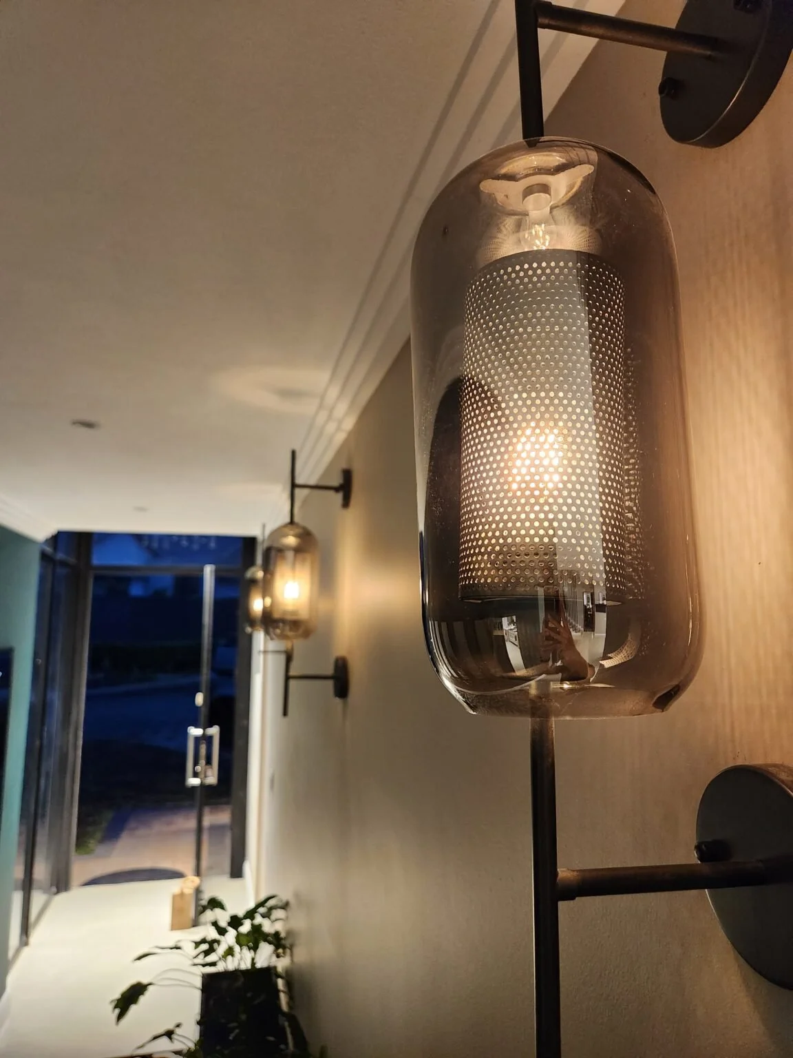 Close-up of a modern wall sconce light fixture with a cylindrical glass cover and metallic base mounted on a wall, with similar fixtures visible further down the wall.