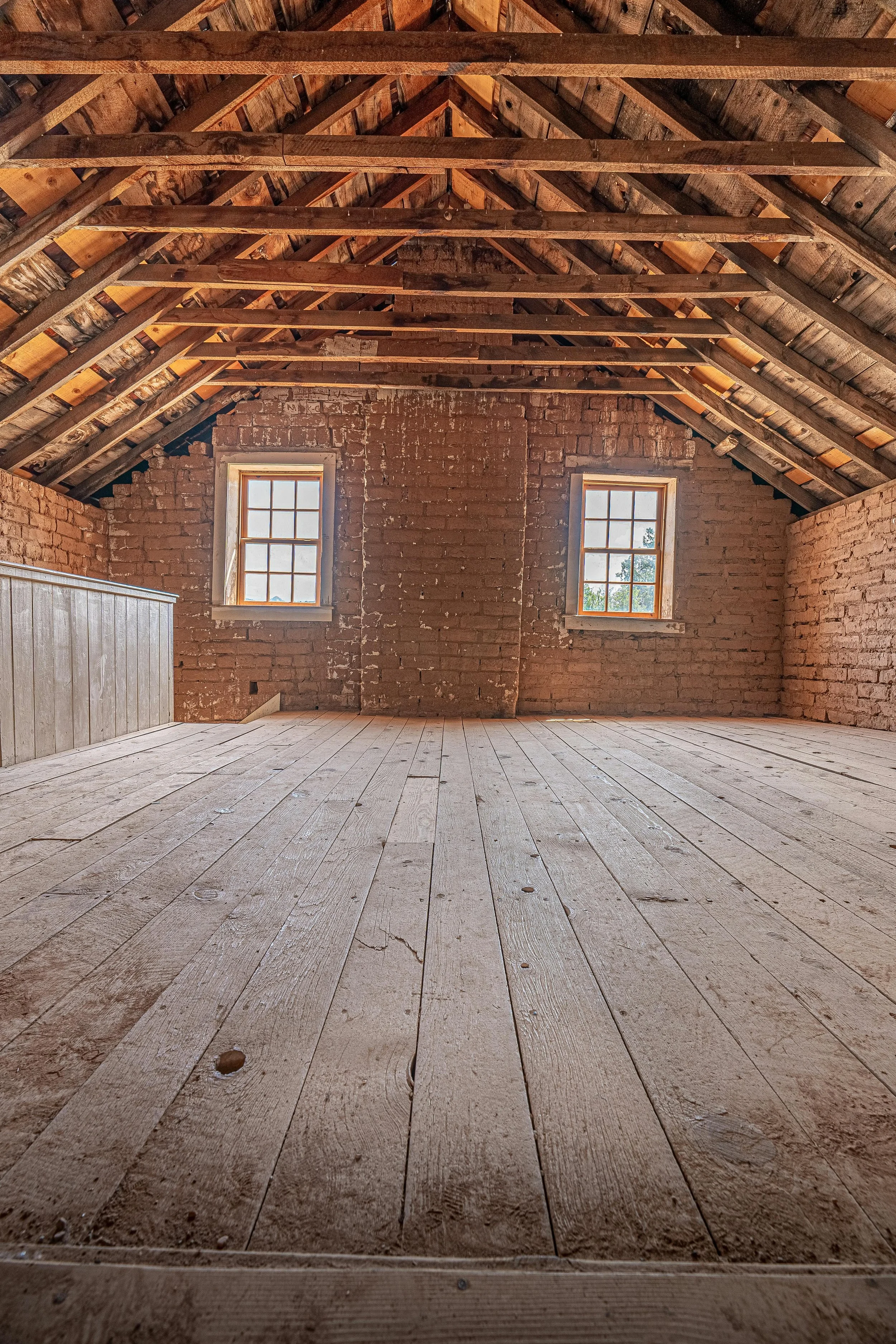 Unfinished attic with exposed wooden rafters, wood flooring, brick walls, a brick chimney, and 2 windows on the back wall.
