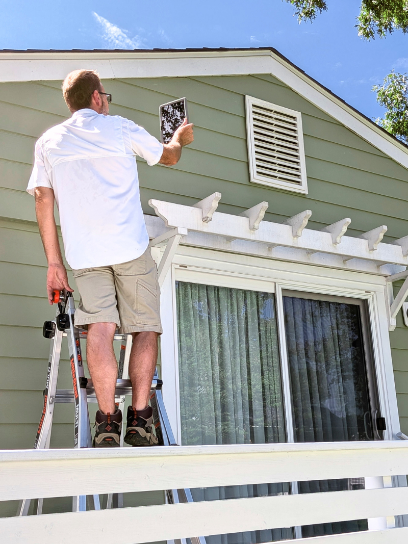 Inspector photographing attic vent from ladder with tablet.
