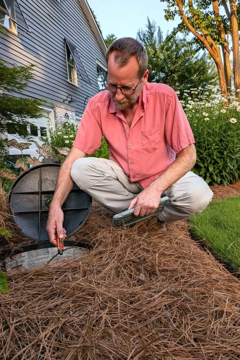Shawn DePenning squatting in a bed of pine straw, testing an inground propane tank for a gas leak while holding an inspection tablet.