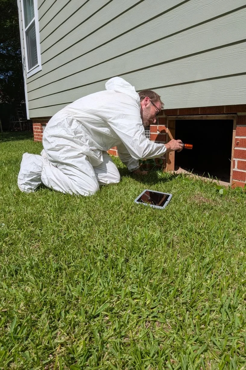 Shawn inspecting a crawlspace on his knees in protective coveralls, shining a flashlight into the brick foundation opening with his tablet on the grass nearby.