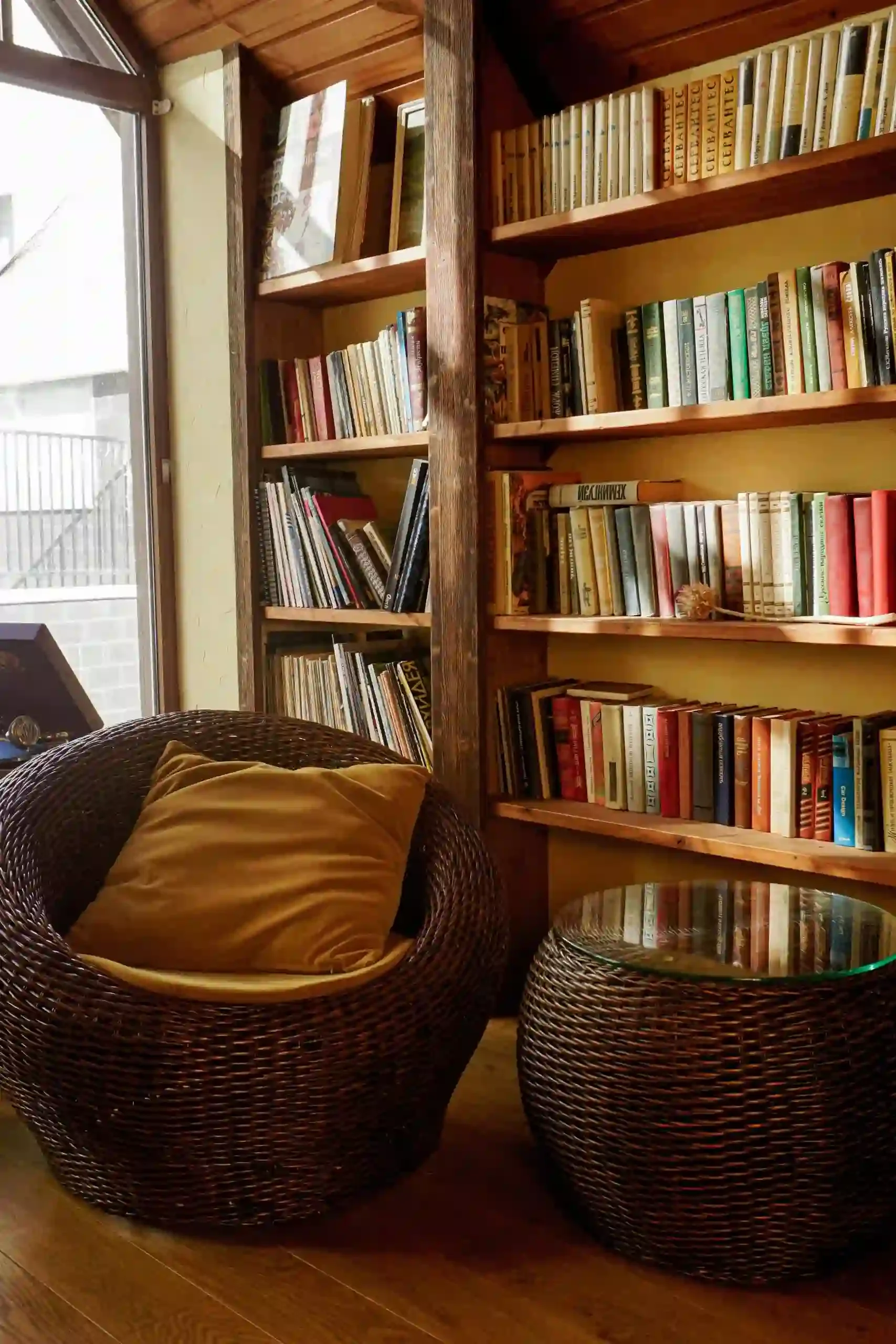 Cozy reading nook with wicker chair, warm wood bookshelves, and a glass-top table beside a large window.