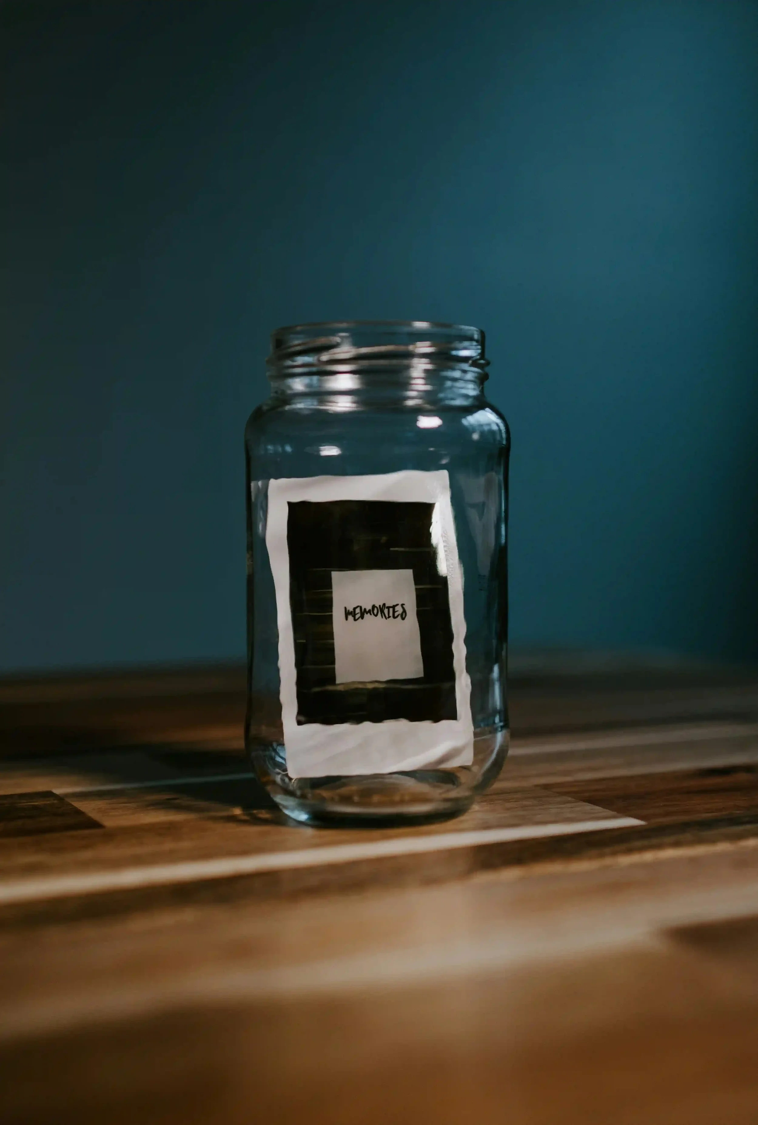 Glass jar labeled “Memories” resting on a wooden table against a dark blue background.