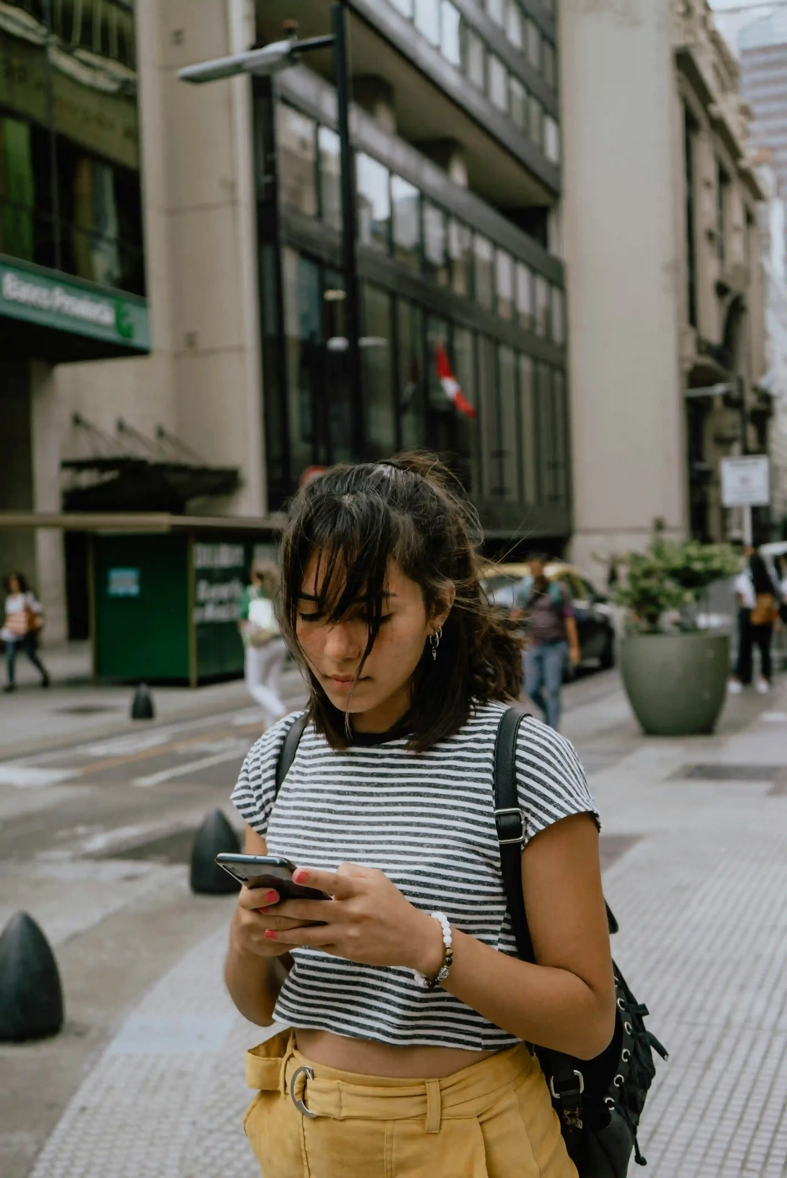 woman with wind-blown hair looking down with focused expression at her phone while walking on a city sidewalk