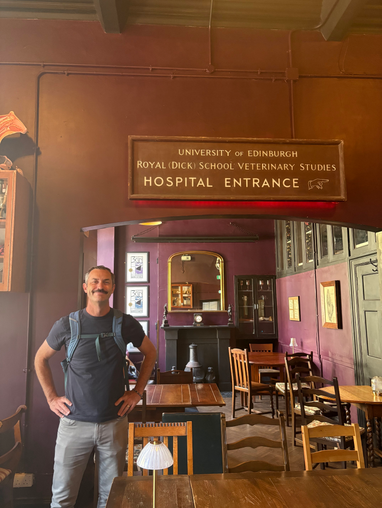 A man with a backpack standing in a vintage-style room inside a building, with a sign for the University of Edinburgh Royal Dick School of Veterinary Studies indicating a hospital entrance.