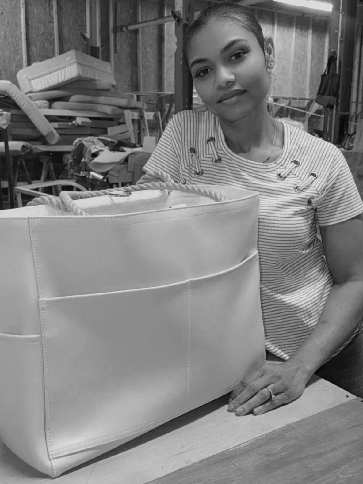 A woman sitting at a table in a workshop, with a large white handbag in front of her. The workshop has shelves with foam and other materials in the background.