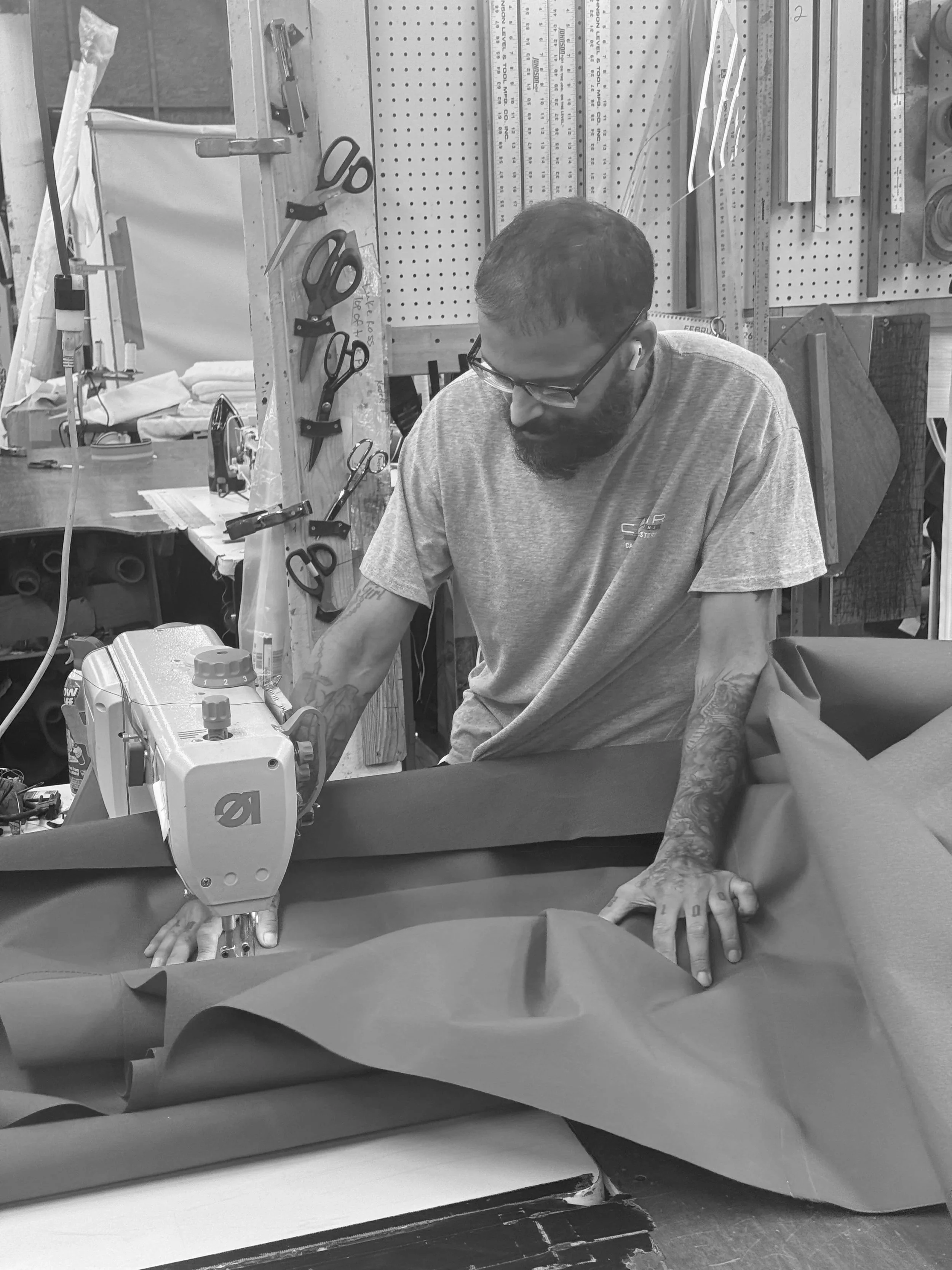 A man working in a workshop or sewing room, operating a sewing machine. He is wearing glasses and wireless earbuds, with tattoos visible on his left arm. The background includes scissors, rulers, and other tools on a pegboard.