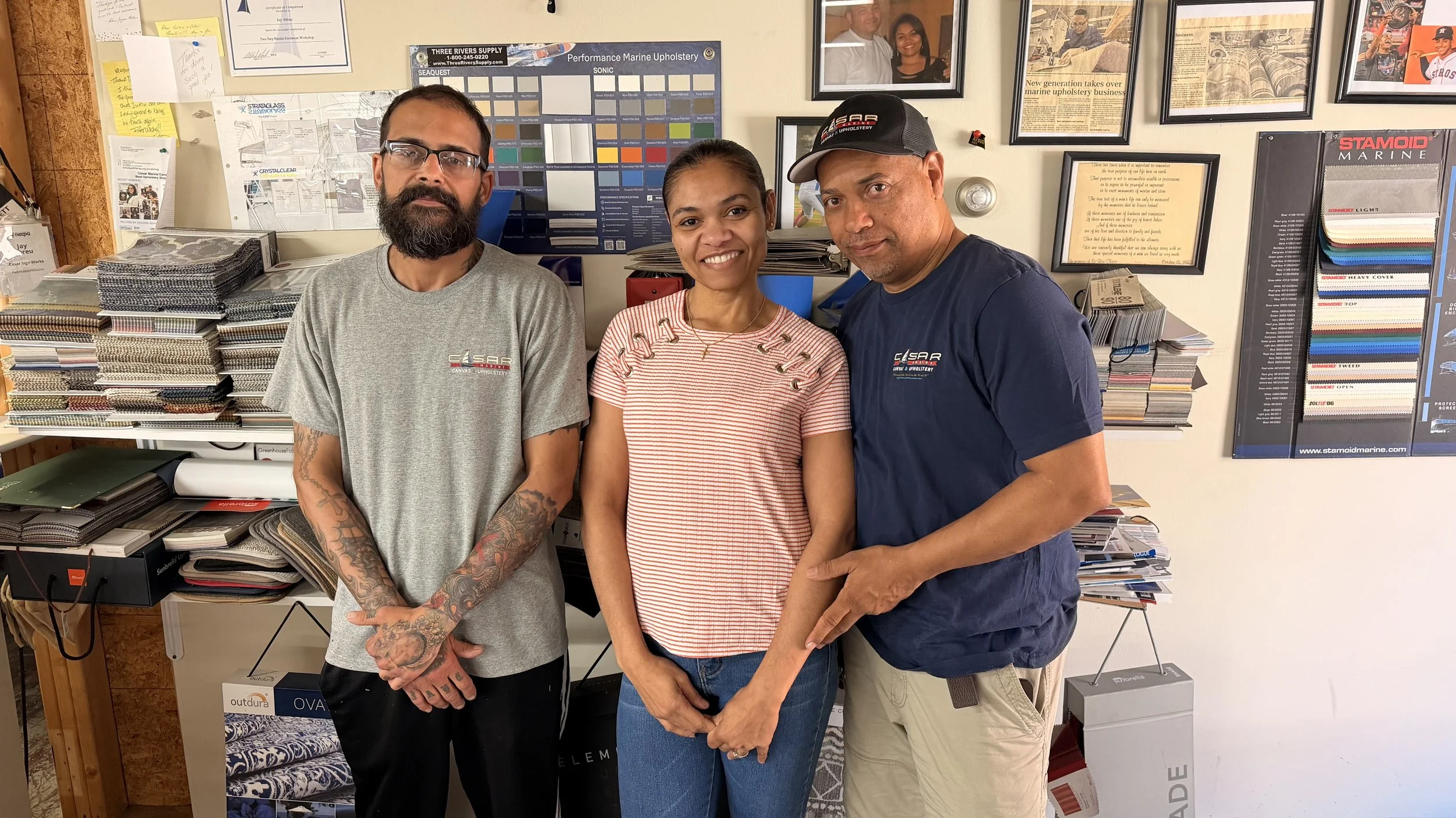 Three people standing inside a store or office, surrounded by sample swatches, framed pictures, and various paperwork on the walls. The person on the left is a bearded man wearing glasses and a gray T-shirt with tattoos on his arms. The woman in the middle is smiling, wearing a red and white striped shirt. The man on the right is wearing a baseball cap and a dark blue T-shirt, with his arm around the woman.