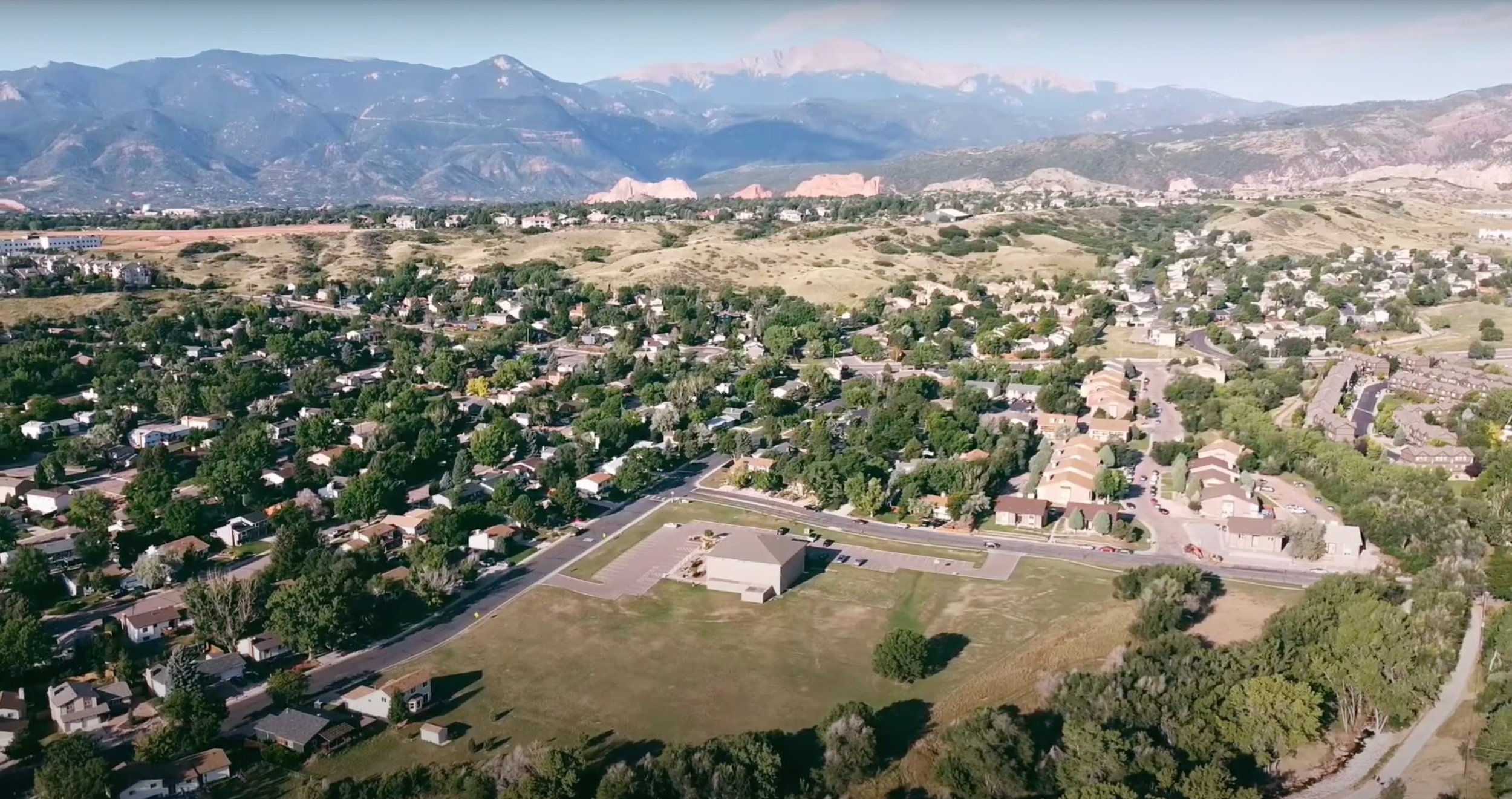 Aerial view of a suburban neighborhood with houses and trees, surrounded by rolling hills and mountains in the background.