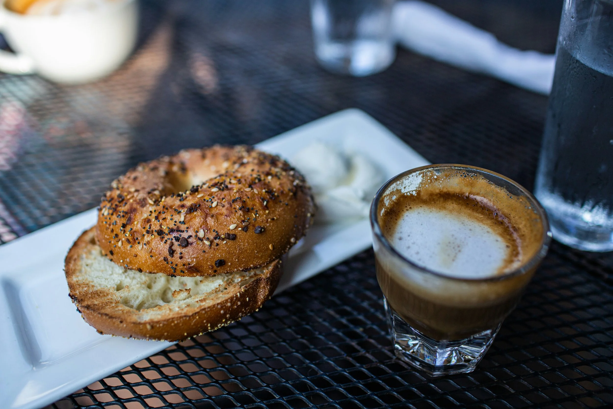 A blueberry bagel with cream cheese on a white plate and two glasses of frothy coffee drink on a black wire mesh table.