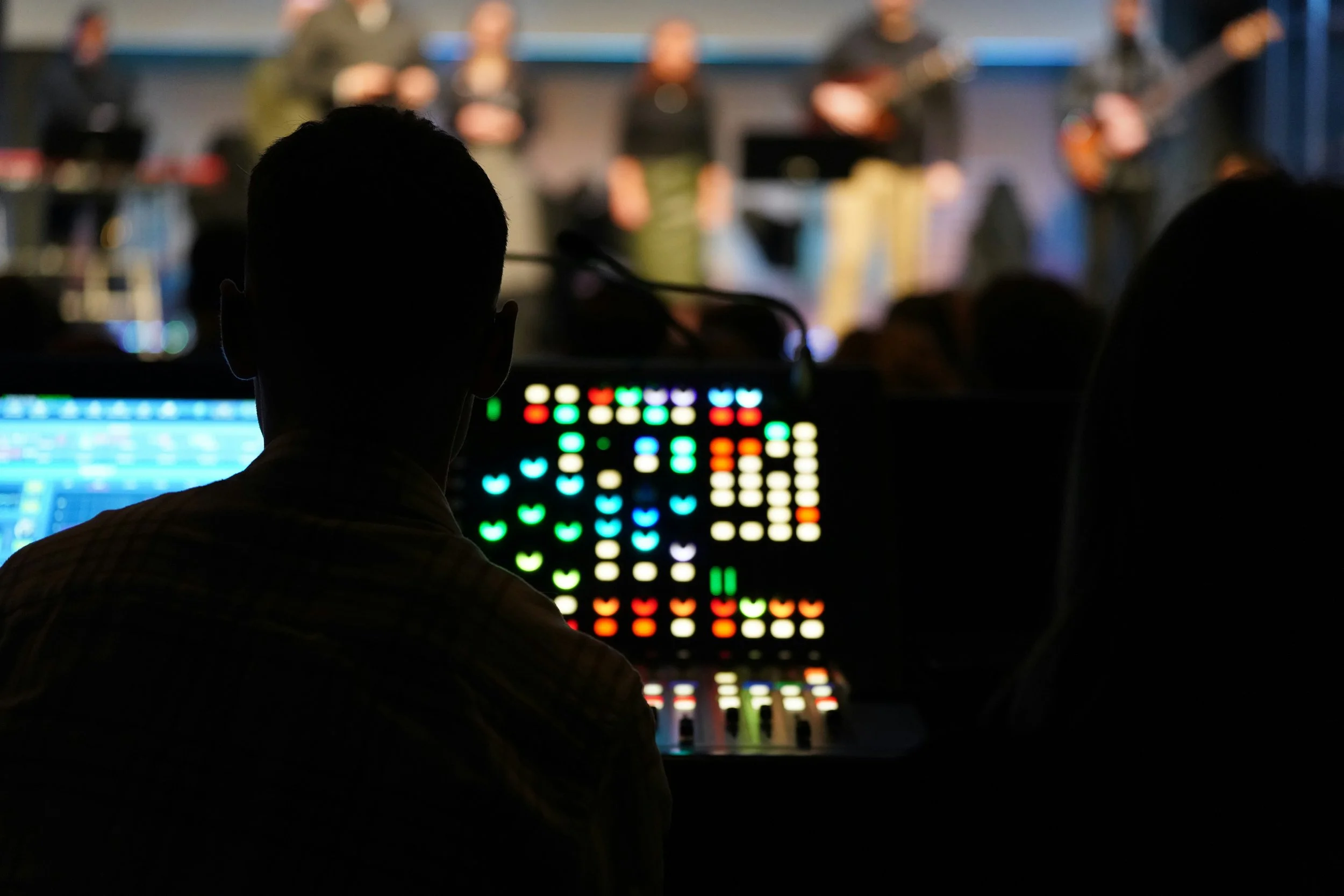 A person working on sound or lighting control equipment in a dark room with a stage and band in the background.