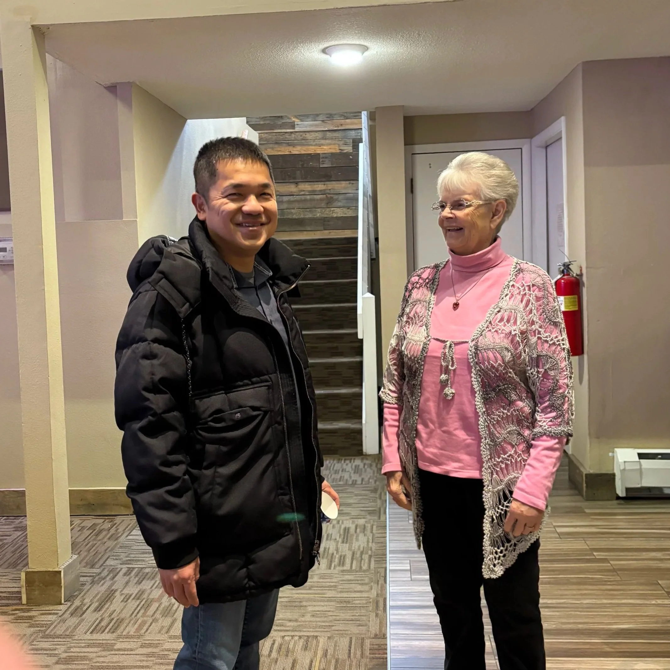 A young man with short dark hair and a dark jacket, smiling, standing next to an elderly woman with white hair and glasses, wearing a pink turtleneck and a patterned cardigan, both indoors, in front of a wooden wall and a mirror.