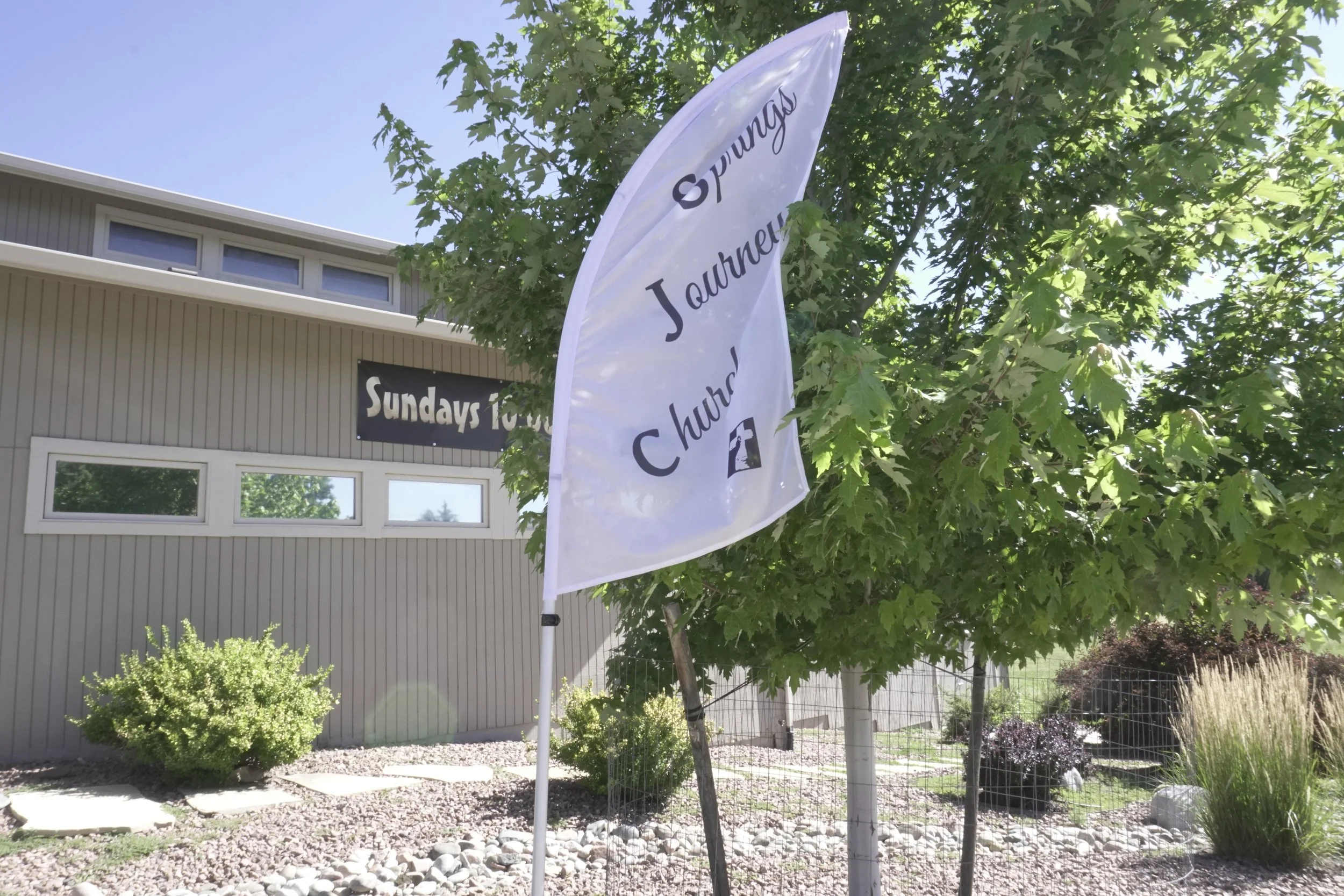 A white flag with black text that reads 'Sundays' and 'Church' is attached to a pole outdoors, surrounded by green trees and bushes, with a gray building in the background and a clear blue sky.