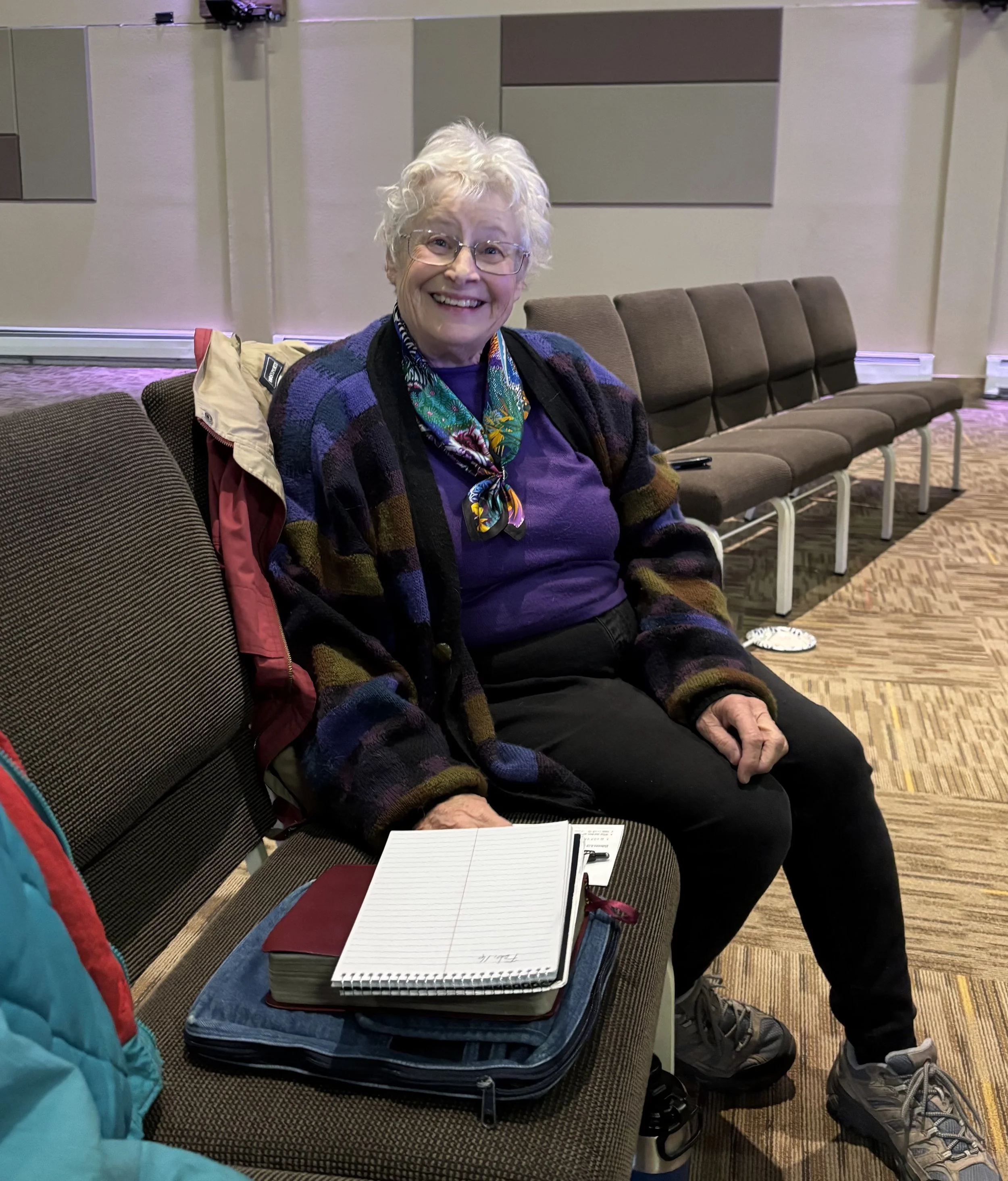 A smiling elderly woman with white curly hair and glasses, sitting on a row of brown chairs in what appears to be an airport or waiting area. She is wearing a colorful scarf and a multicolored cardigan. In front of her, there is an open notebook on top of an open suitcase. To her left, part of a person wearing a blue jacket is visible. There are other empty seats and luggage in the background.