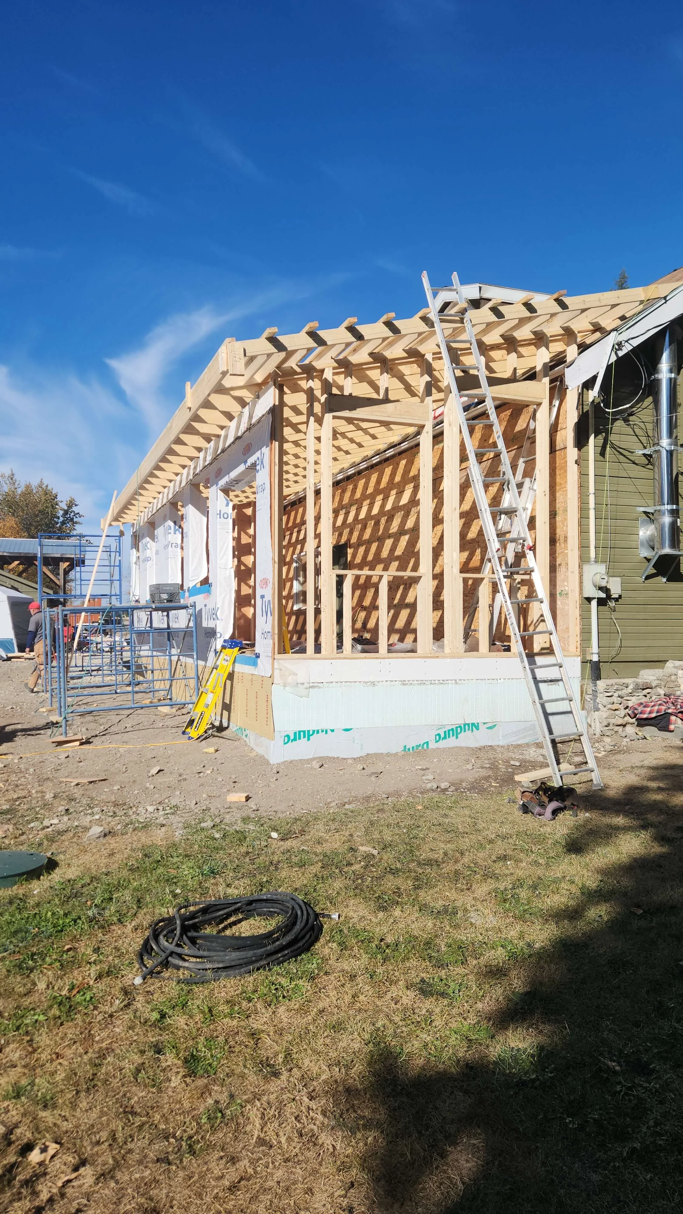 Construction site of a wooden building with a ladder leaning against it, under a clear blue sky.