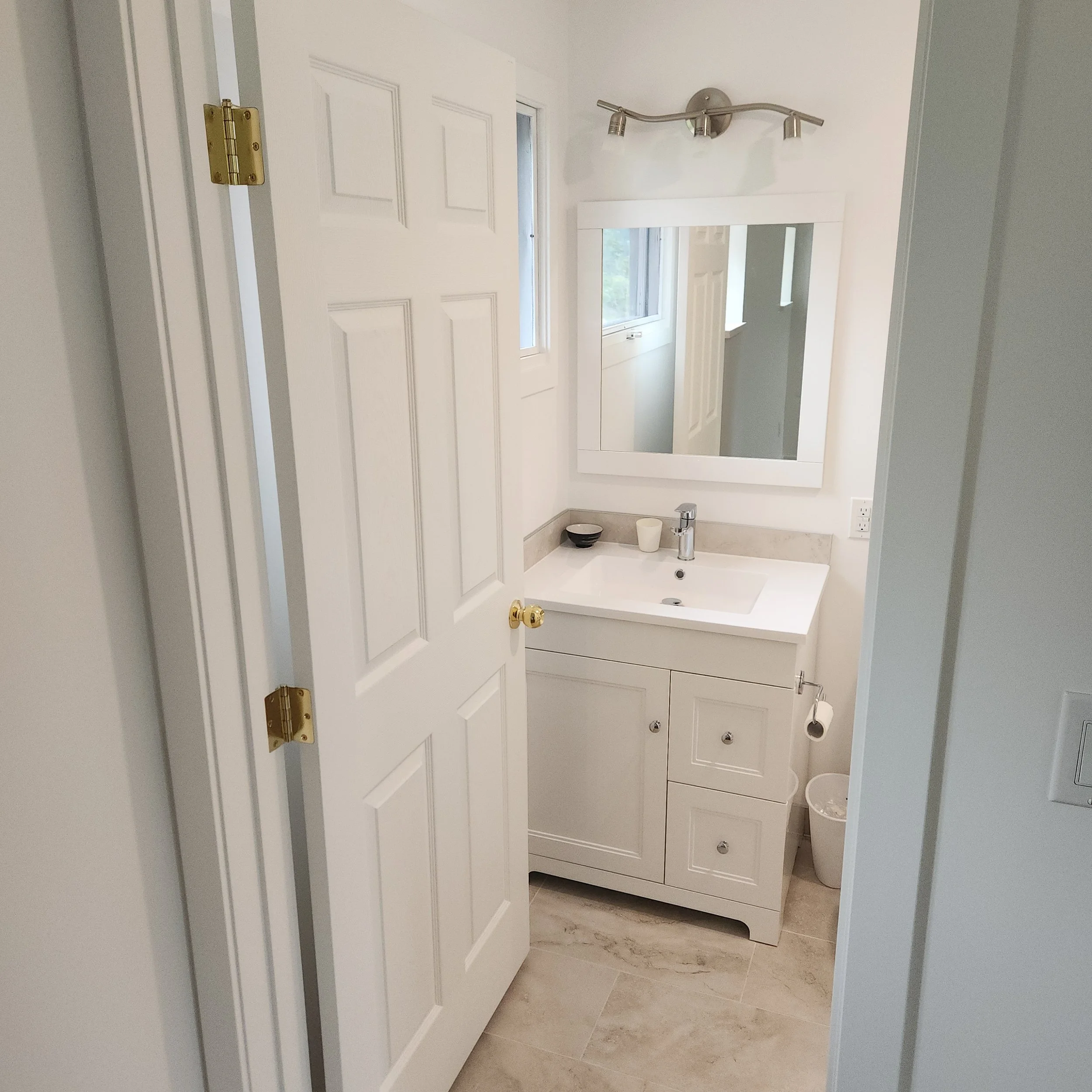 A clean and bright bathroom with a white vanity cabinet, a rectangular mirror, a small window, and a modern wall-mounted light fixture with three adjustable spotlights.