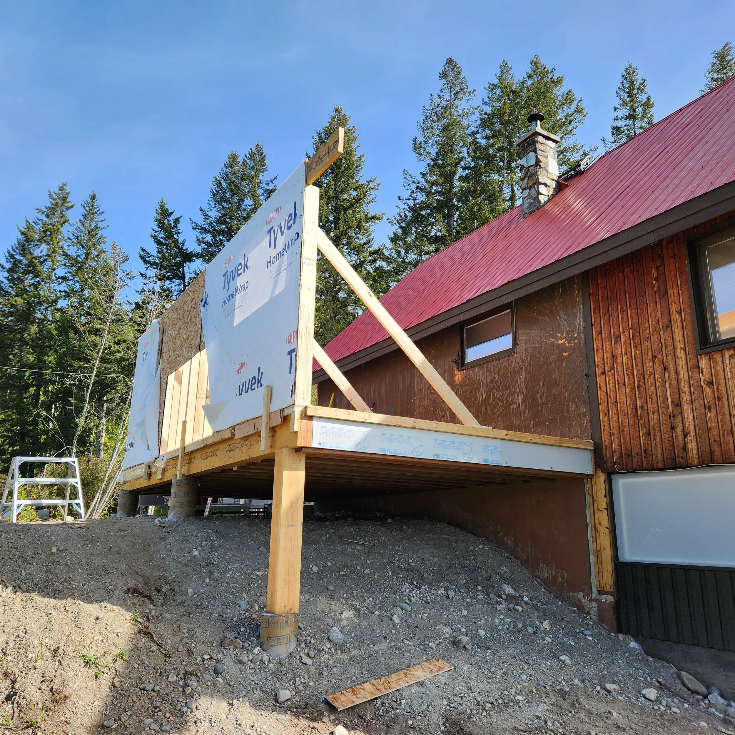 Construction site with a wooden frame and partially built house, a step ladder, a chimney, and a red metal roof, surrounded by trees under a blue sky.