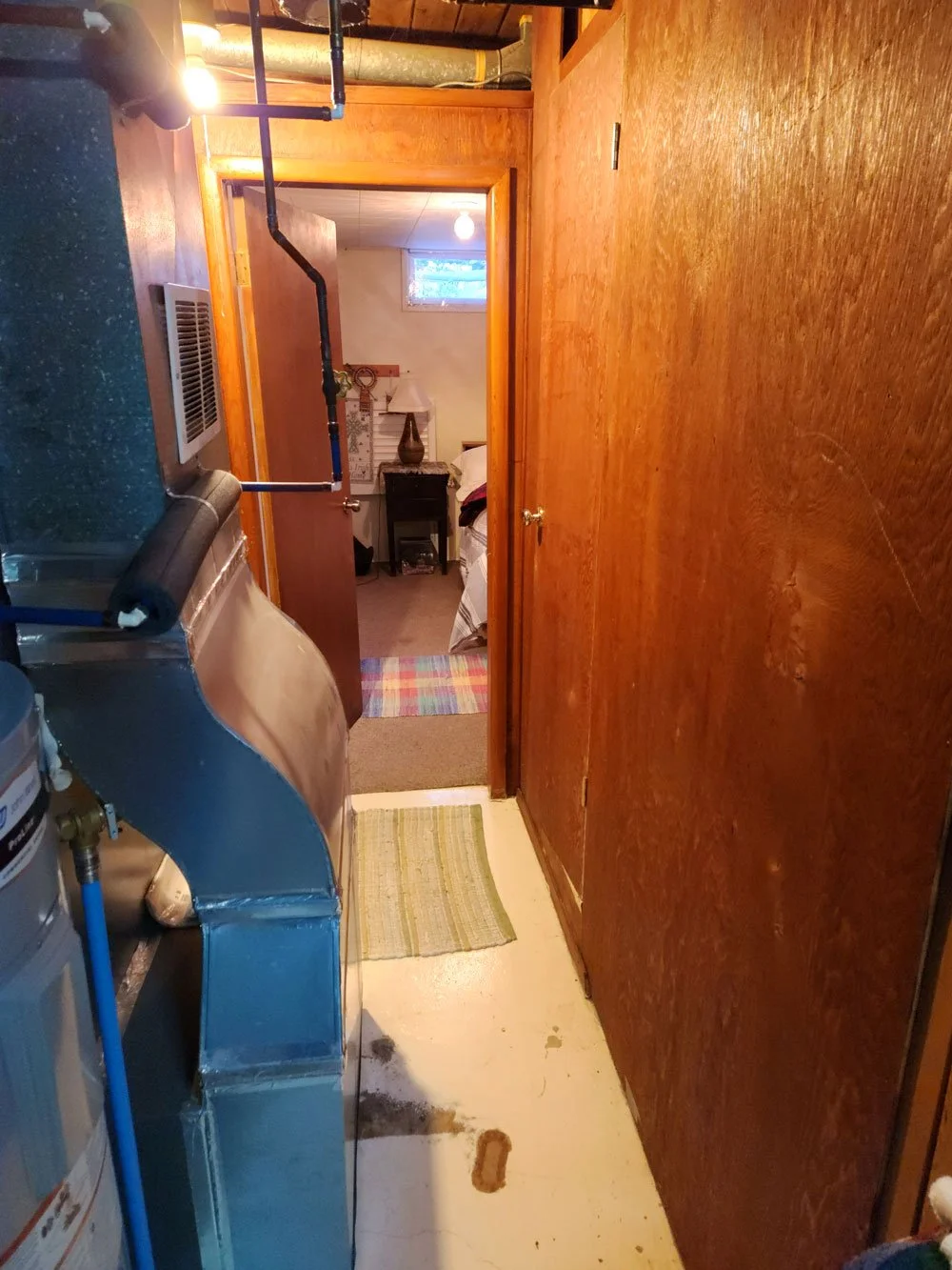 A narrow laundry or utility room with wood-paneled walls and a concrete floor. To the left, there is laundry equipment including a washing machine and dryer, with pipes running along the top. The room leads into a bedroom with a window, a bed, a smal