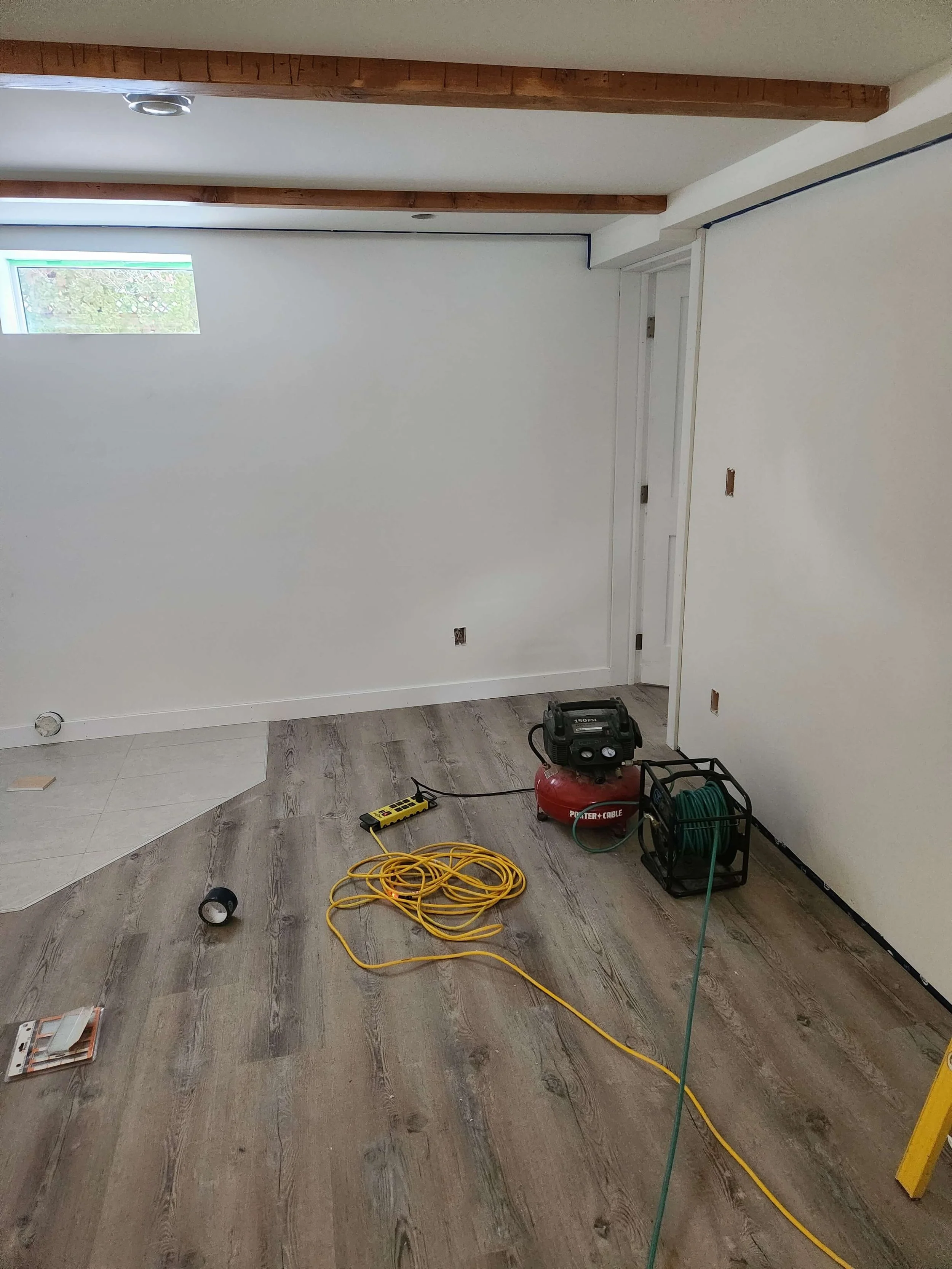 A room under renovation with unfinished walls, tools, and equipment such as a compressor, extension cord, and measuring tape scattered on the floor.