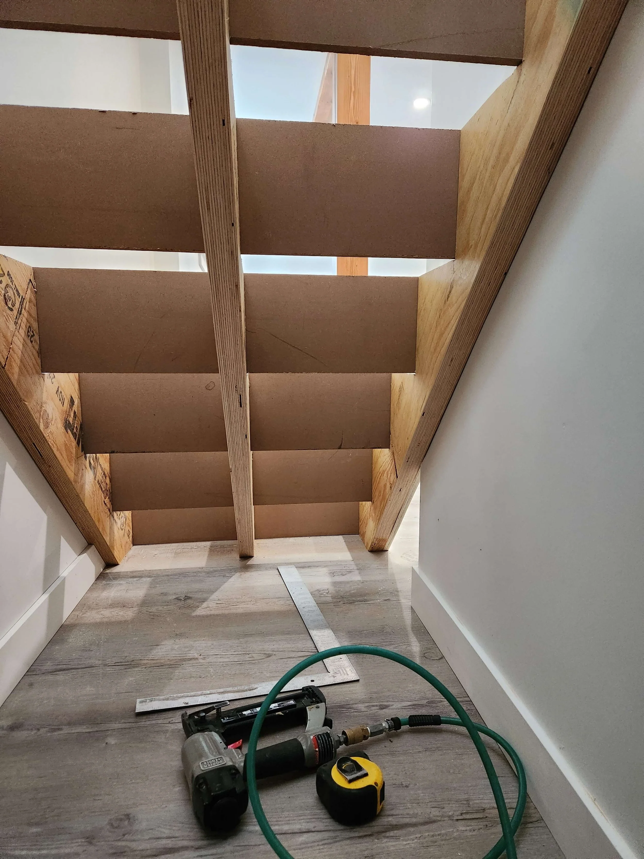 Partially assembled wooden furniture frame on a light-colored wooden floor, with tools and measuring tape around it, and a ruler on the floor.
