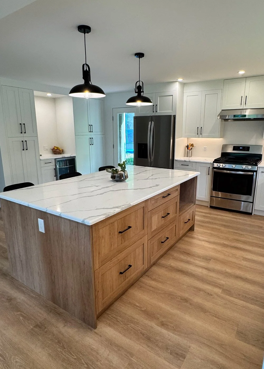 Modern kitchen with a large island featuring a white marble countertop, wooden base with drawers, and black handles. Stainless steel fridge and oven, white cabinetry, and pendant lights hanging over the island. Wooden floor and some small plants on t