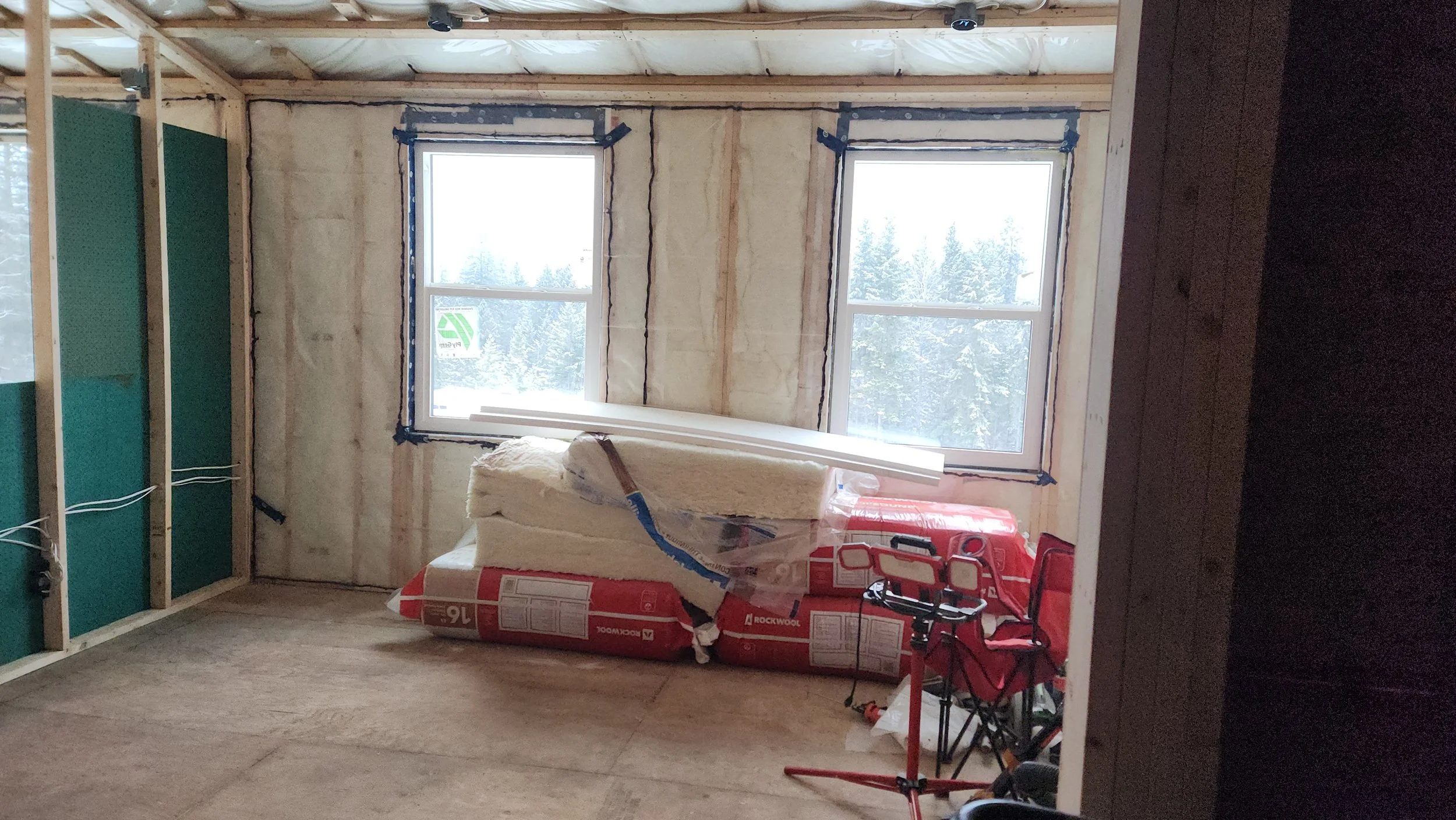 The image shows an unfinished room with two large windows. Construction materials, including insulation and boxed drywall, are in the room. The walls are partially framed with wooden studs, and some wiring is visible. Outside, snow-covered trees can 