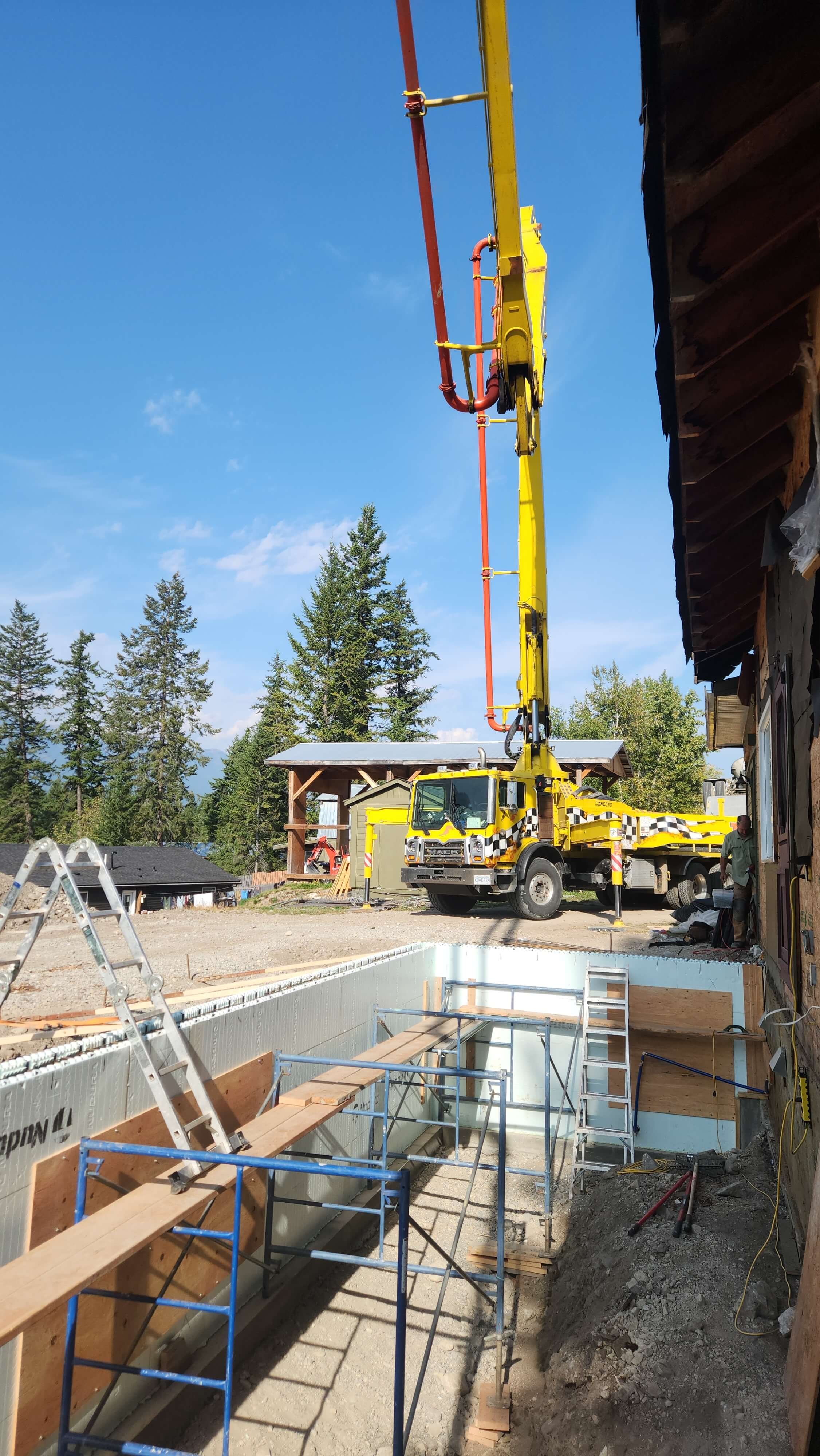 Construction site with a large yellow crane truck lifting objects, wooden scaffolding, and building framework, set against a backdrop of trees and a blue sky.