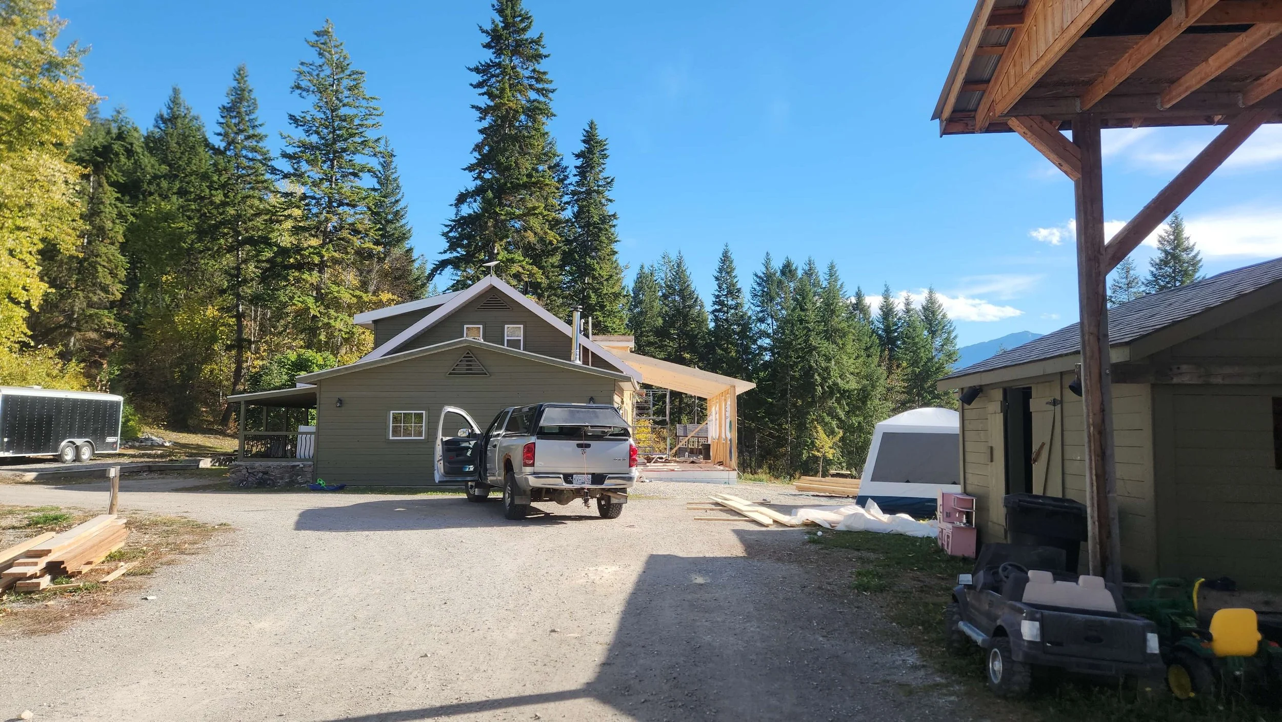 Construction site with two houses, one painted green, with trucks and building materials around, surrounded by tall pine trees and mountains in the background.