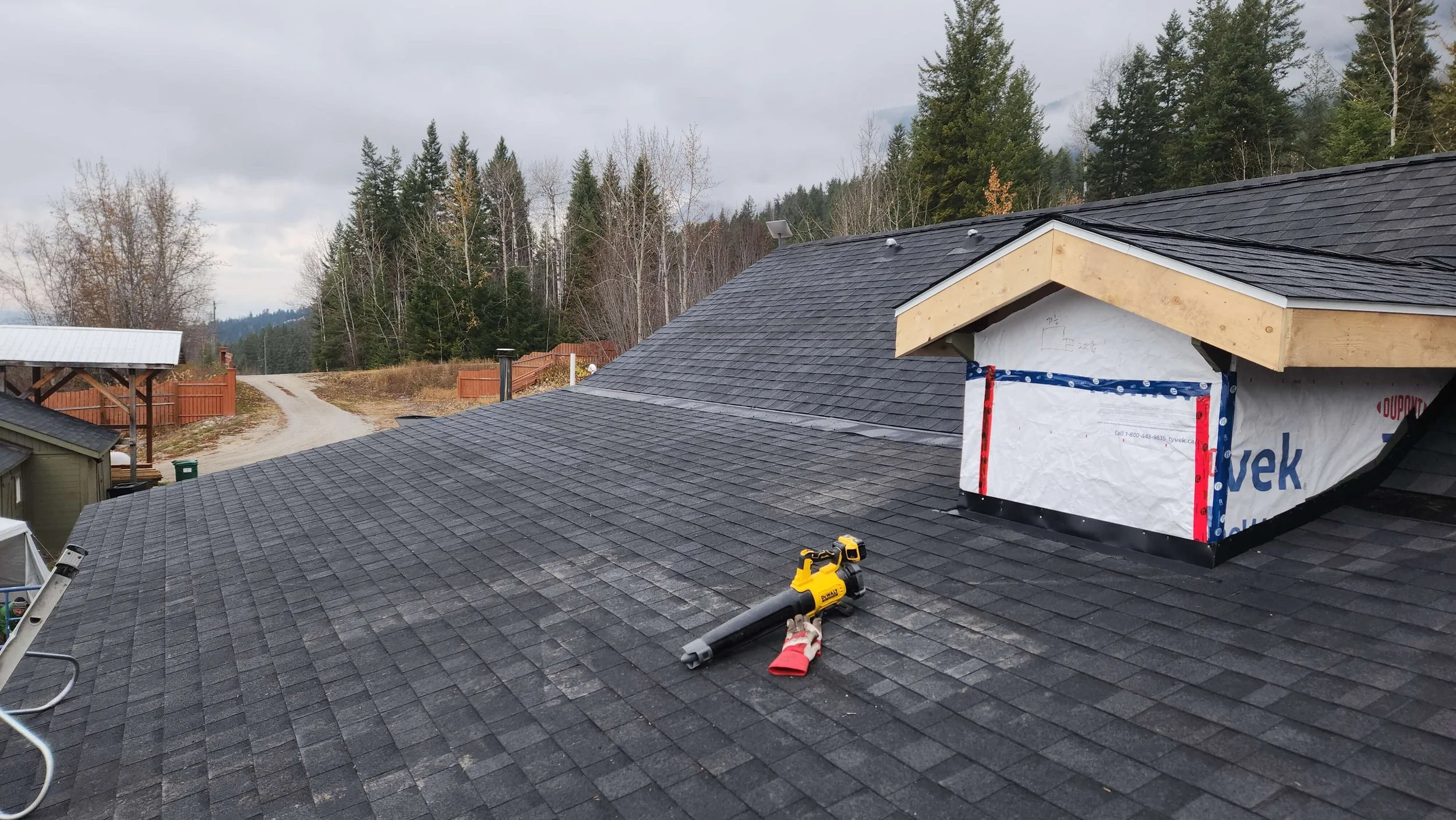 Residential house roof under construction with new black shingles, tools on the roof, and a partially completed dormer area with exposed wood and house wrap, surrounded by a rural landscape with trees and a dirt road.