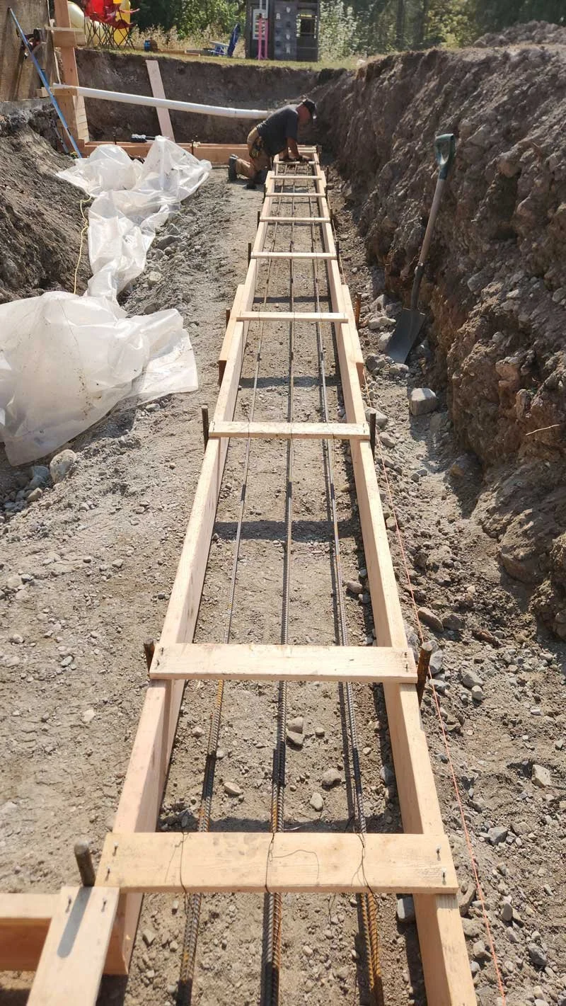 Worker installing rebar in a wooden framework for concrete foundation in an excavation site, with construction tools and plastic sheeting nearby.