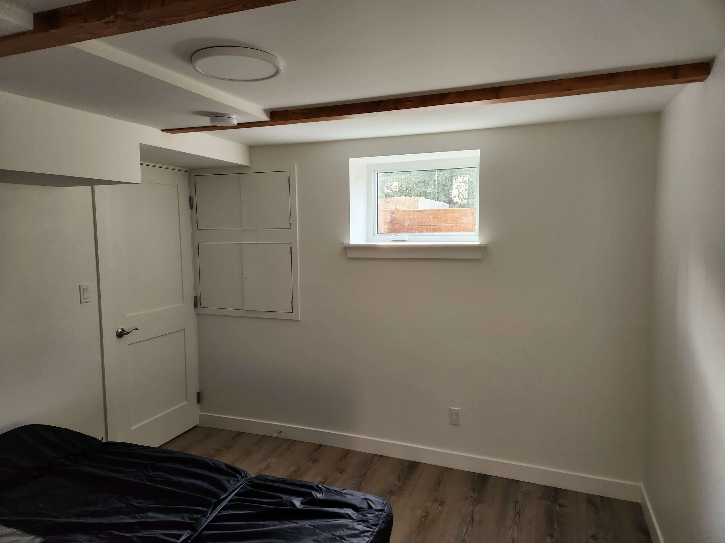 Empty white room with a window, closed white door, ceiling light, smoke detector, and wooden beams.