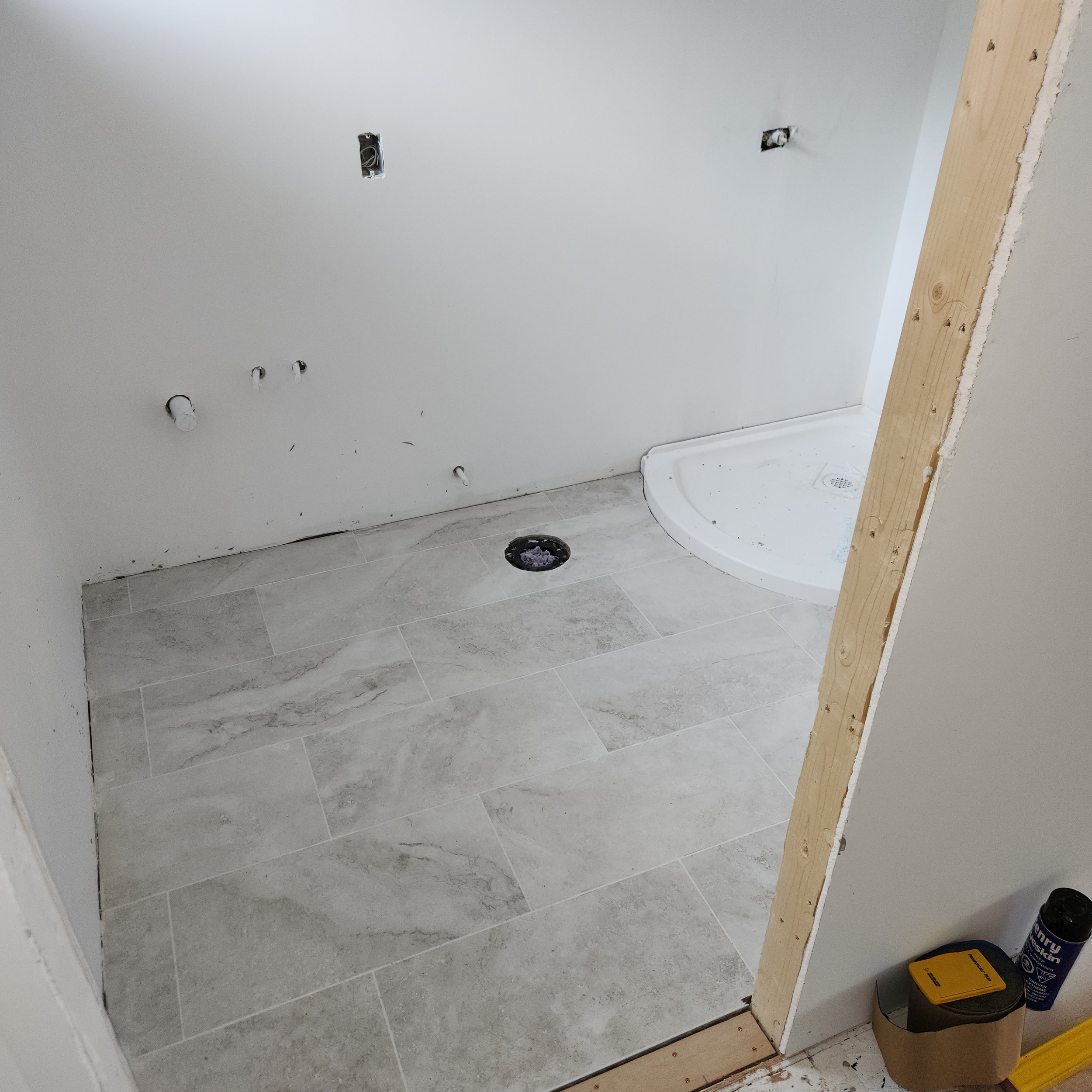 A bathroom under construction with gray tile flooring, white walls, and exposed electrical wiring. There are multiple holes in the wall for fixtures, and a wooden support is partially installed. Construction supplies, including a yellow level and a c