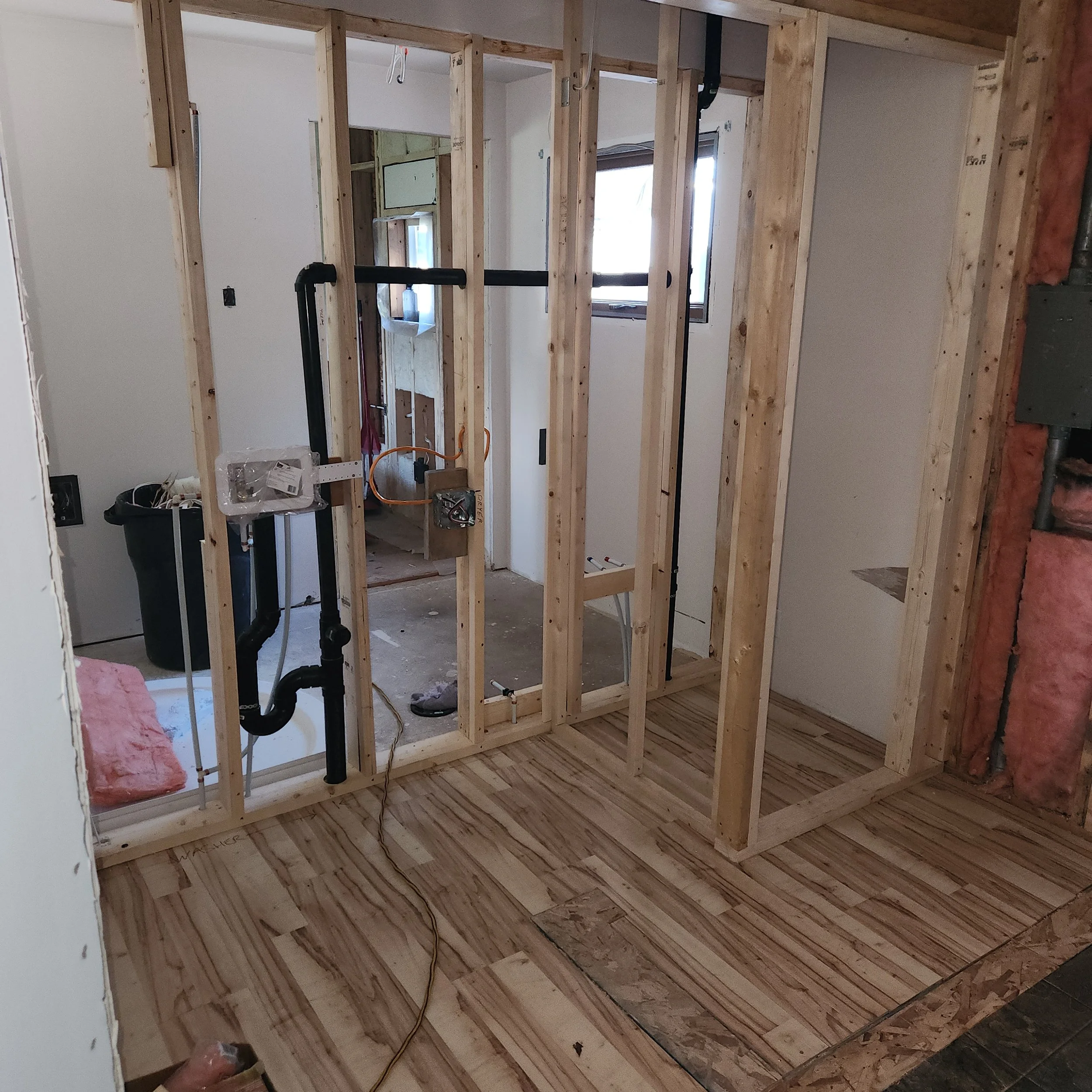 An unfinished room under construction with exposed wooden framing, plumbing pipes, and some electrical wiring visible.
