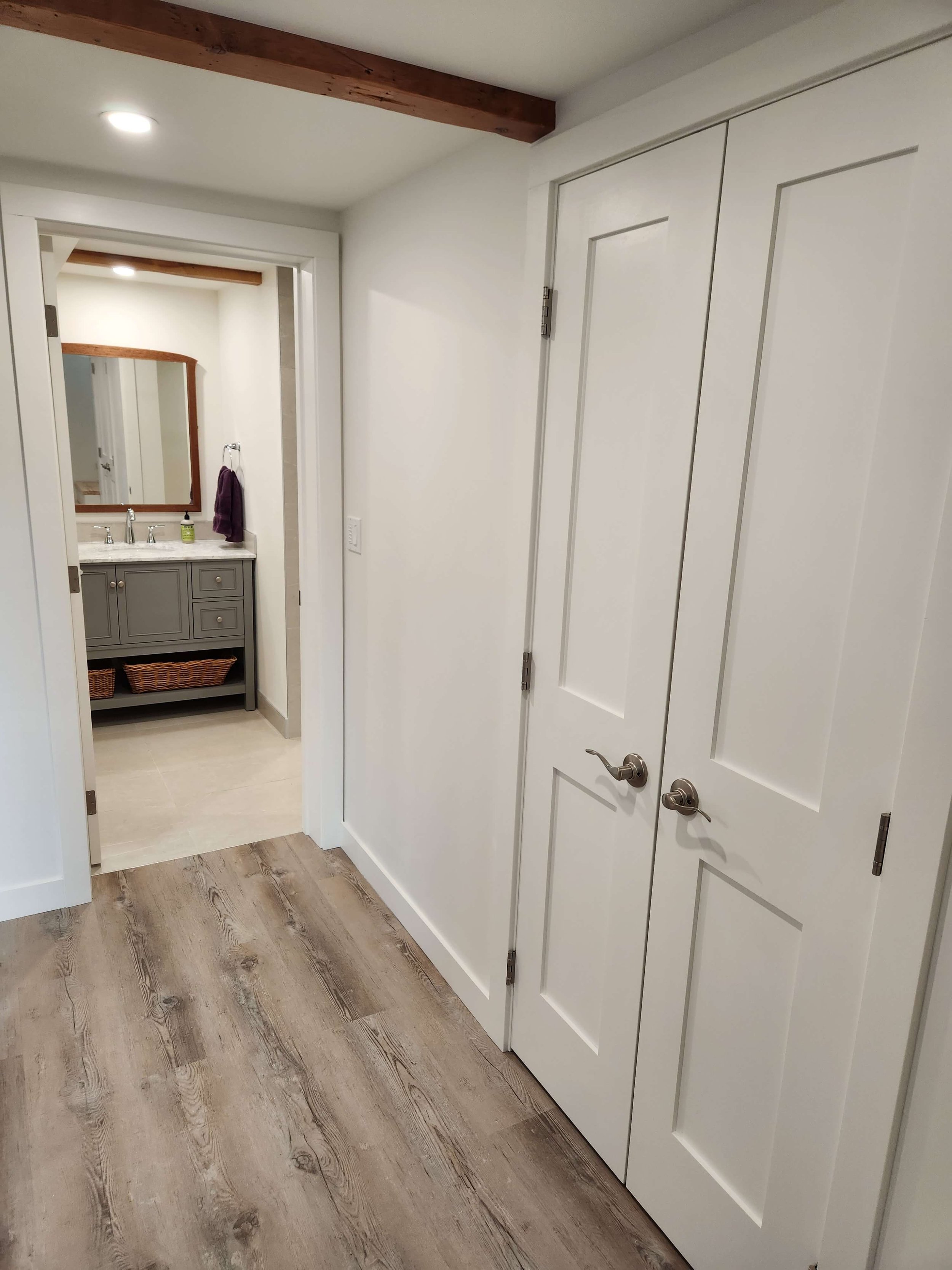 A hallway leading to a bathroom with a gray vanity, mirror, purple towel, and soap dispenser. The hallway has wooden flooring and white walls with double doors.