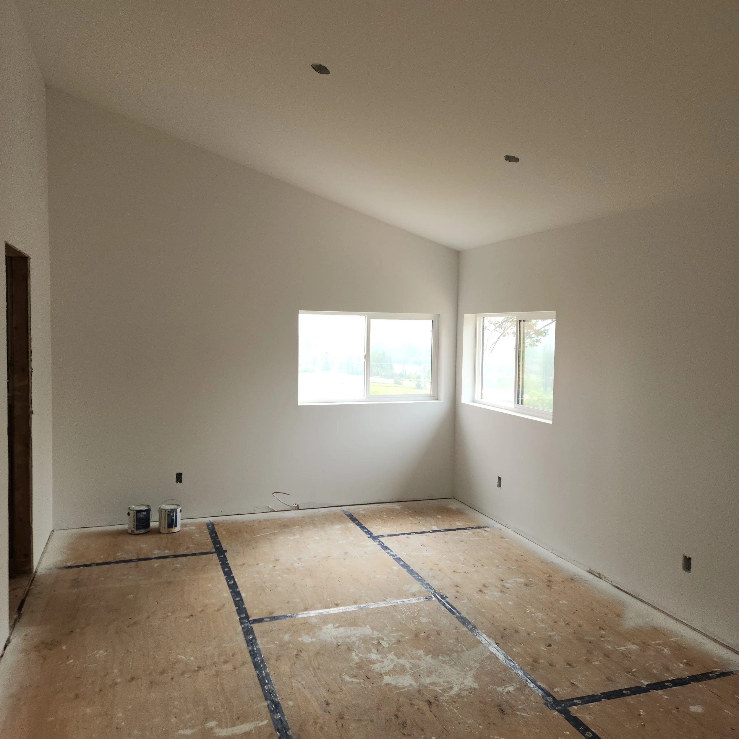 Interior of a room under construction with bare floors, painted walls, and two large windows, with paint cans on the floor.
