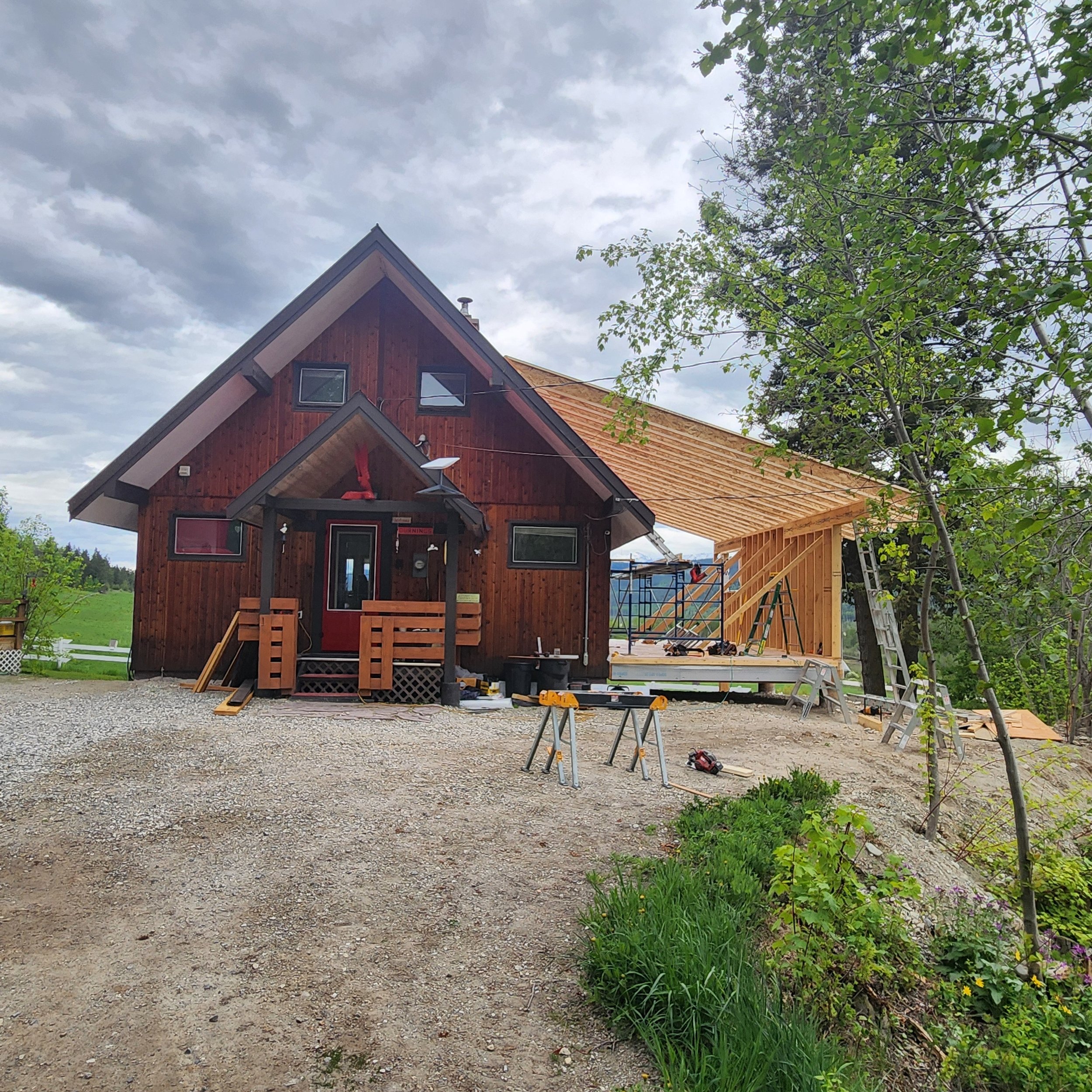A house under construction with a steep roof, surrounded by trees and a cloudy sky.