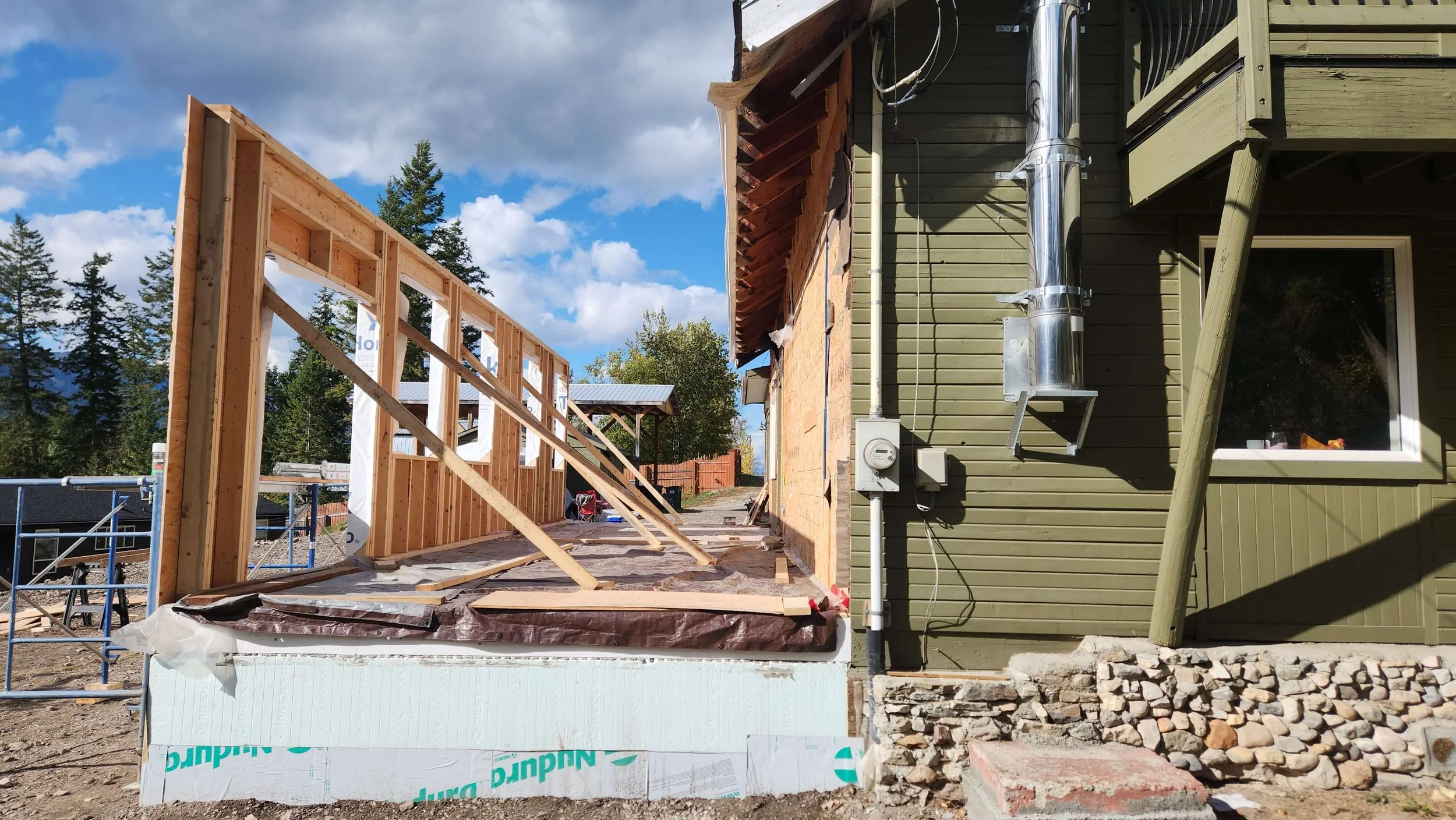 Construction site showing the framework of a new addition being built onto a house, with wooden studs and partial exterior wall.