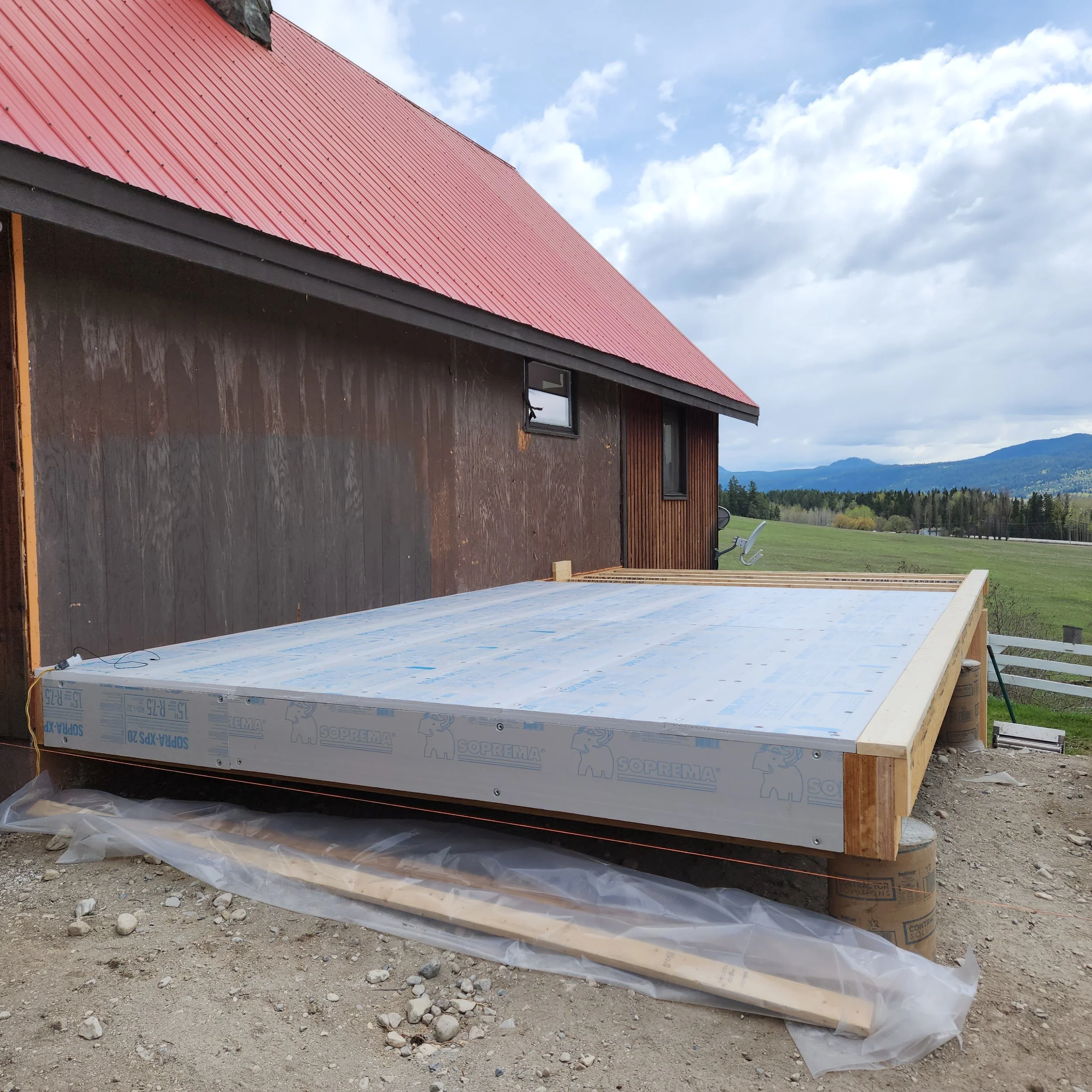 A house under construction with a red metal roof and dark wooden exterior walls and a window, situated in a rural landscape with mountains in the background, cloudy sky, and construction materials on the ground.