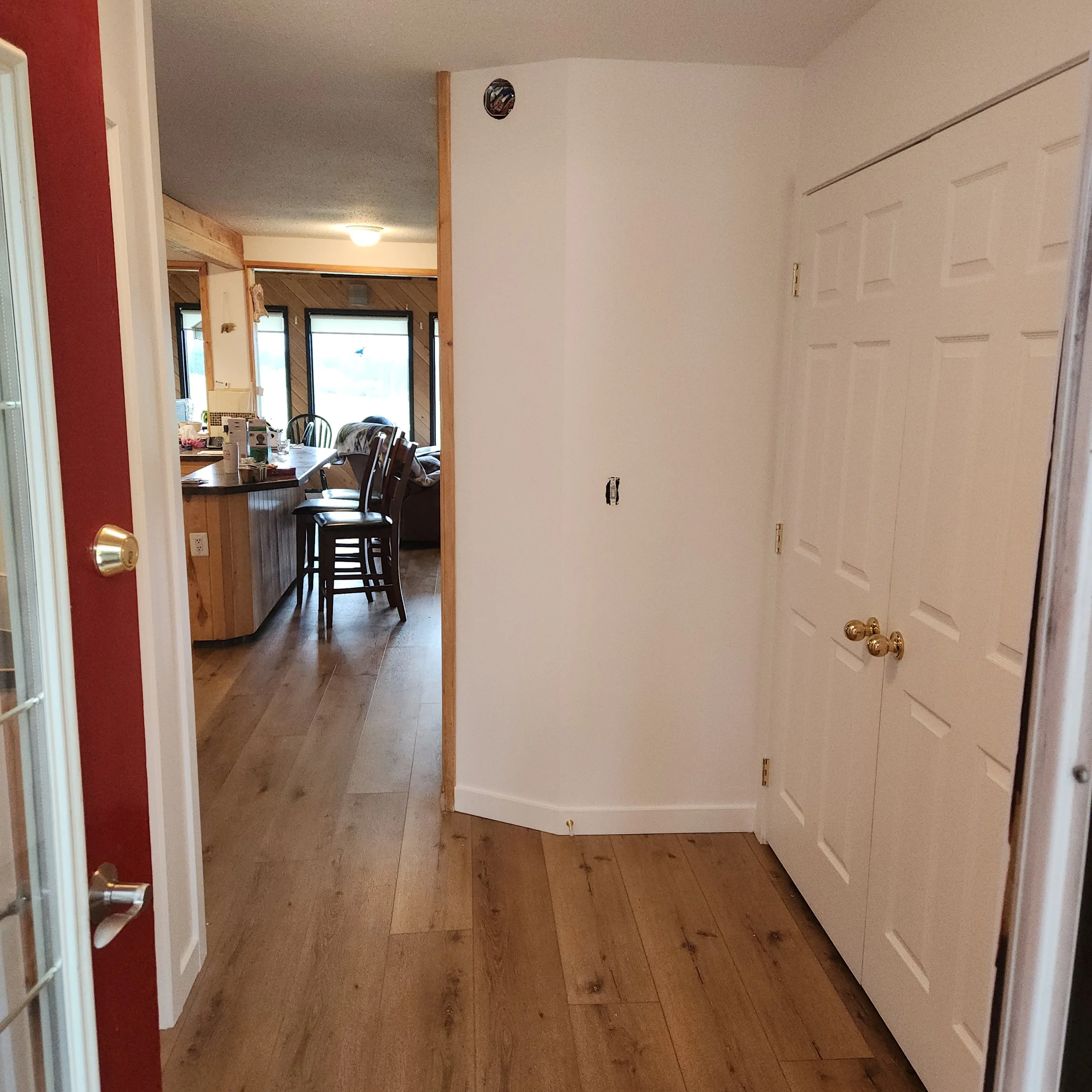 View of a kitchen area seen through an open doorway from a corridor, with a dining table and chairs, and windows with blinds in the background.