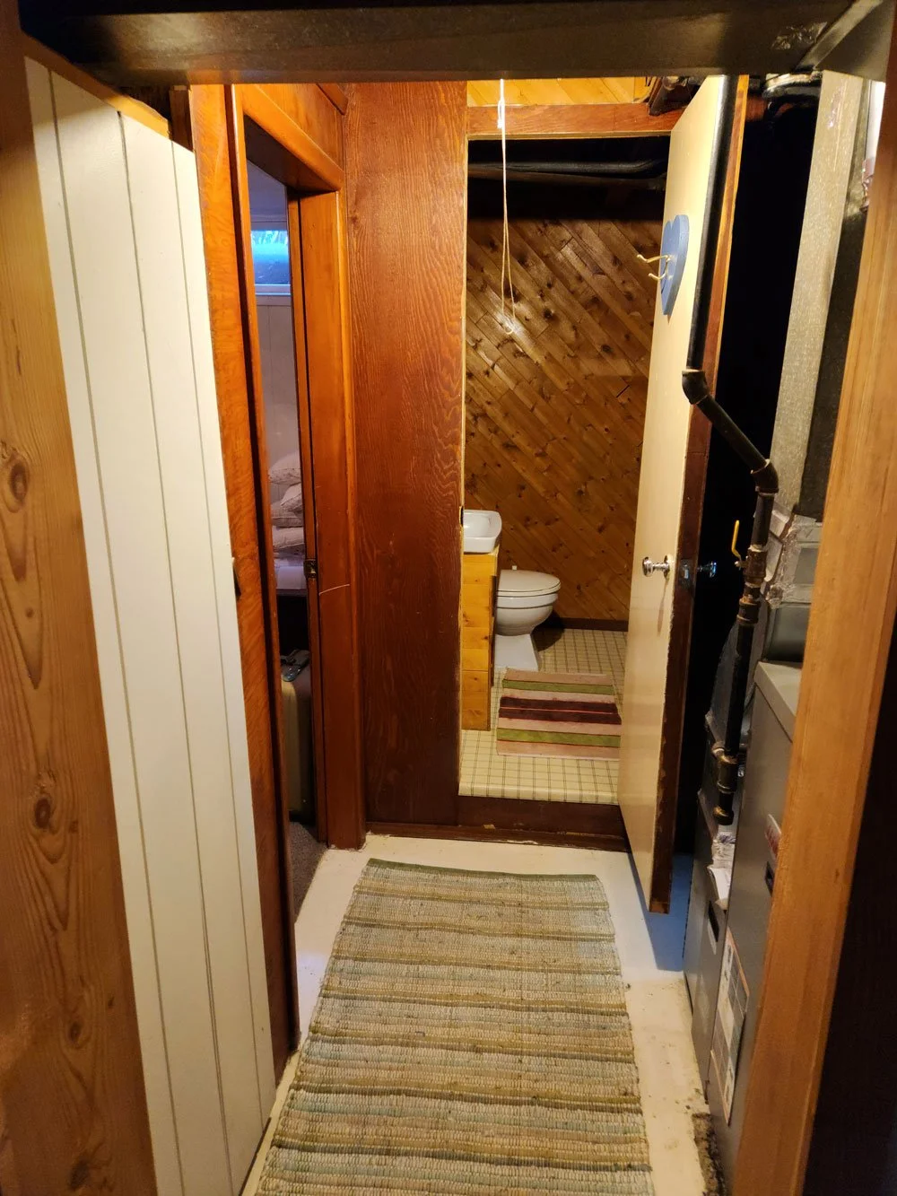 A narrow hallway leading to a small bathroom with wooden walls and ceiling, a toilet, and a small cabinet, with multicolored striped rugs on the floor.