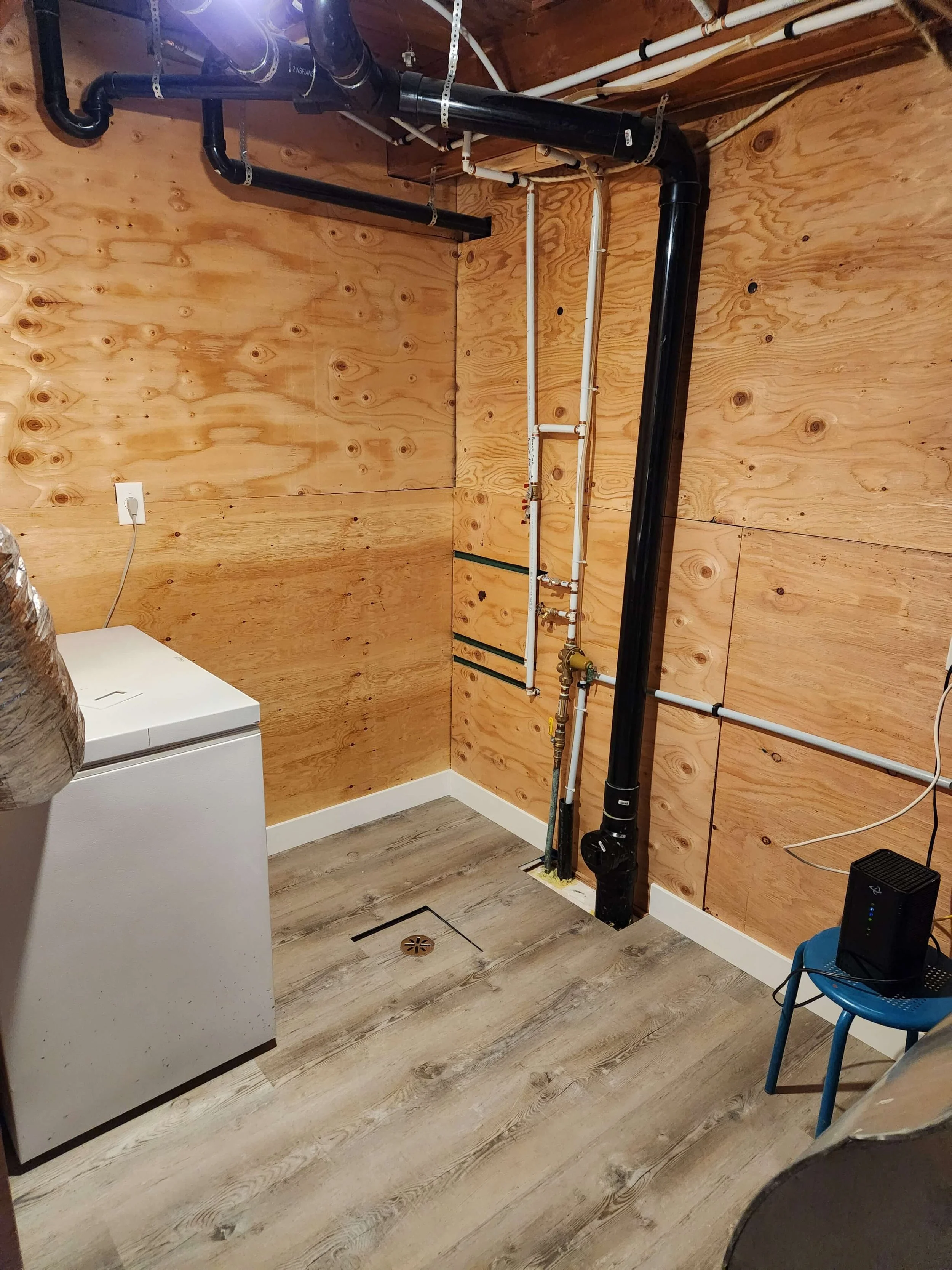 A basement laundry room with wood-paneled walls, a small white washer, black plumbing pipes, white and black plumbing pipes, a blue chair, and a drain in the floor.