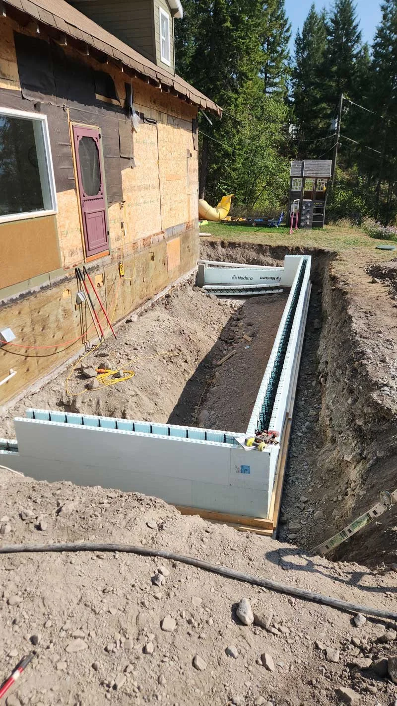 Construction site with a dug trench around a house under renovation, with foundation insulation panels partially installed along the trench.