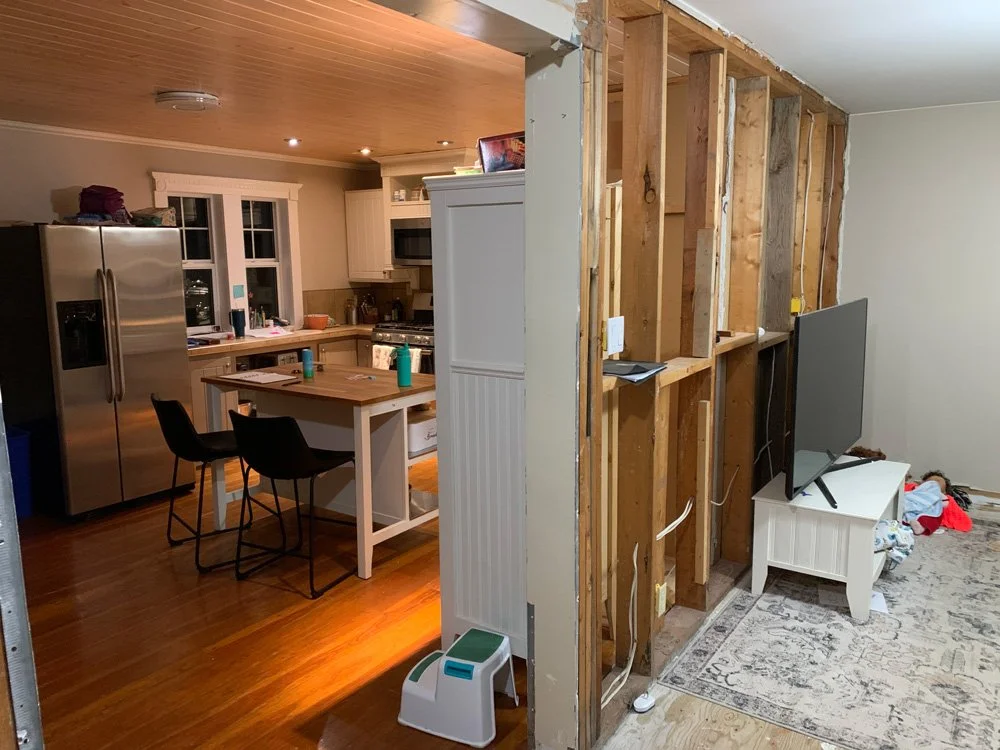 Partially renovated room showing a kitchen with a refrigerator, microwave, stove, and a small table with chairs, separated from a living area with a TV on a white stand by an unfinished wall with exposed wooden studs.