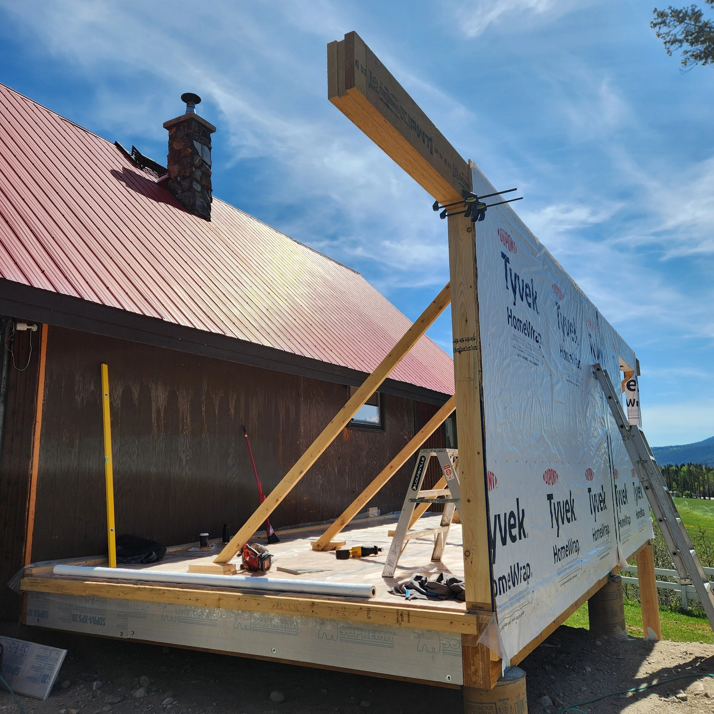 Construction site with a wooden frame for a house, a partially installed chimney on the roof, and a large sign with text and logos, under a partly cloudy sky.