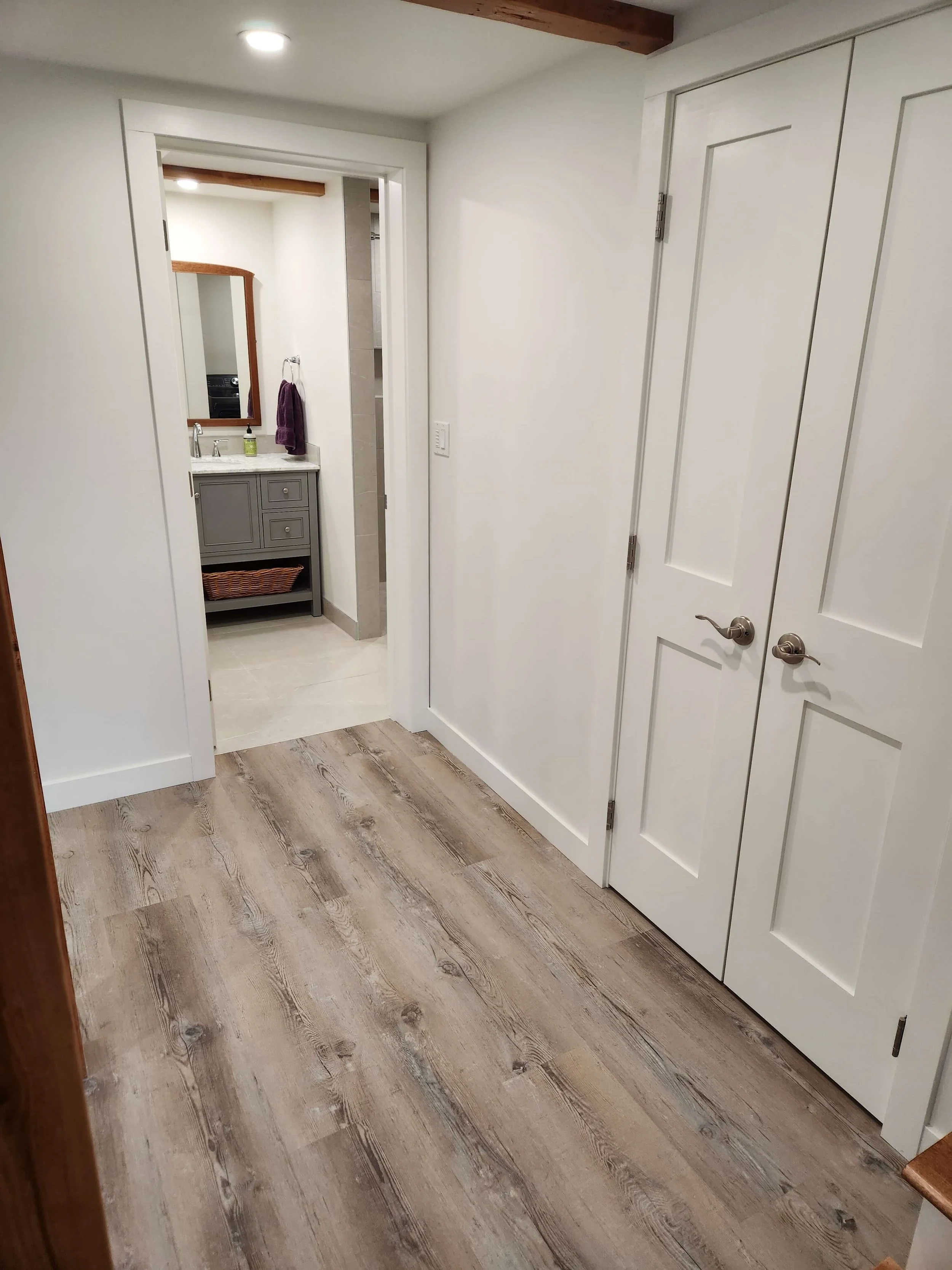A hallway leading to a bathroom with wooden flooring and white walls. The bathroom has a gray vanity with a mirror above it and a purple towel hanging on a ring. There is a partial view of a shower area with beige wall tiles.