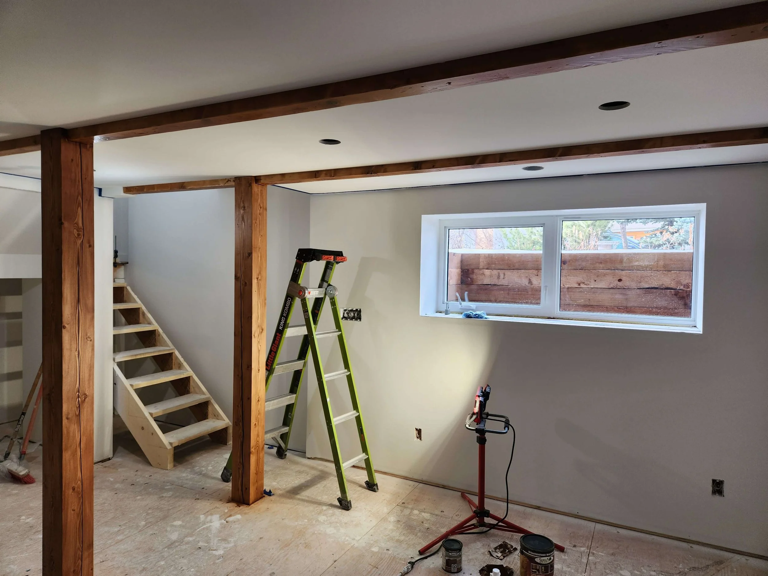 Interior room under renovation with wooden beams, a ladder, stairs, a window, and construction tools and materials.