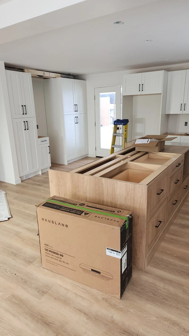 Kitchen under renovation with white cabinets, a cardboard box, and a large wooden island being assembled.