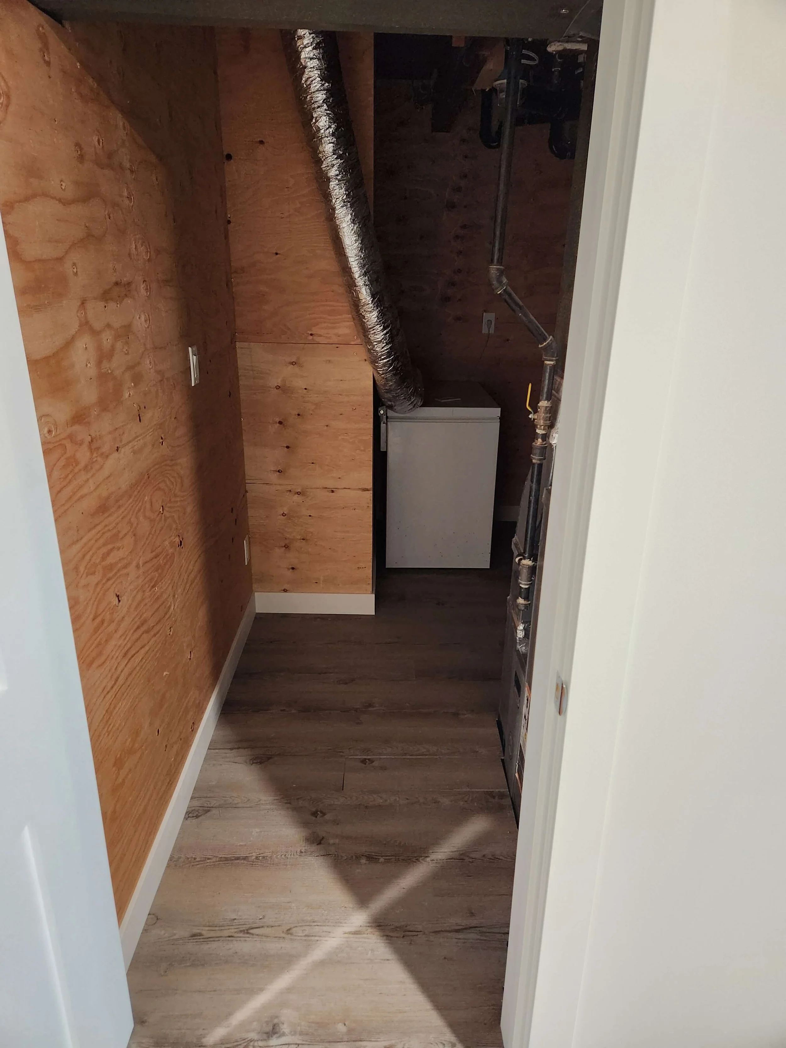 Empty small room or closet with wooden walls, a ventilation duct, a small appliance, and exposed pipes.