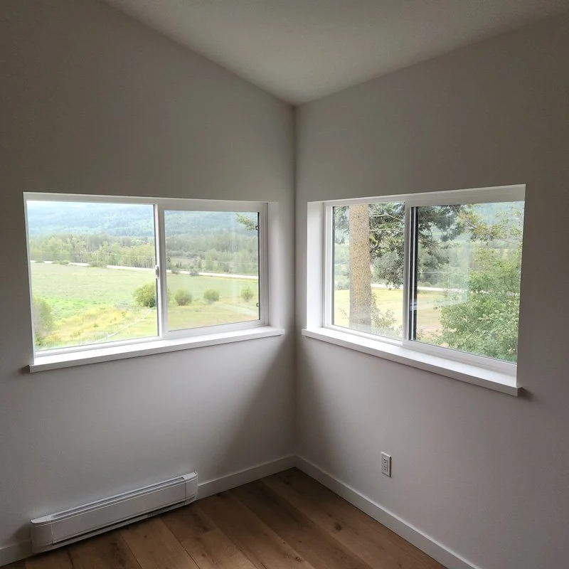 Empty room with two large windows showing a green landscape outside, hardwood floor, and white walls.